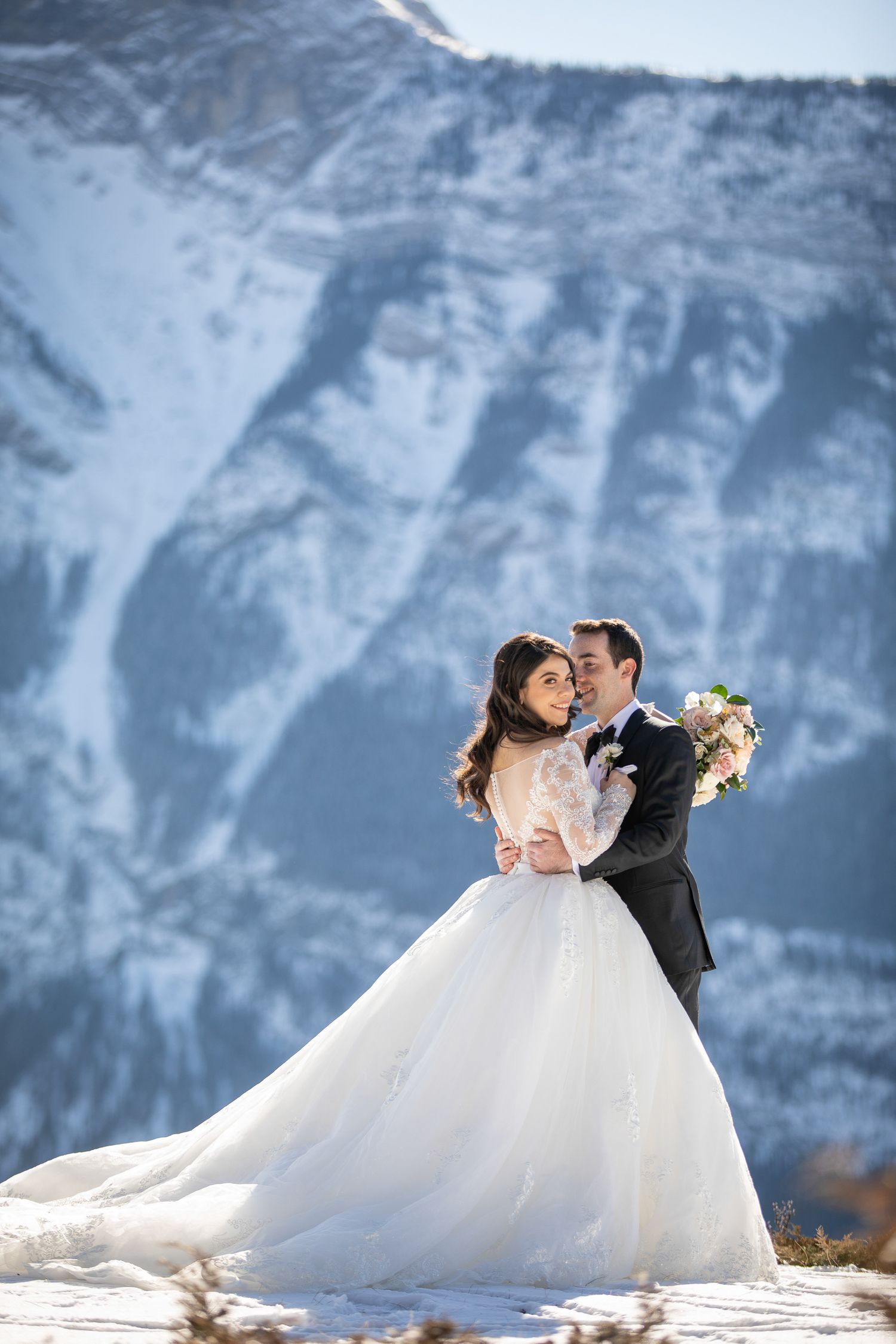 A romantic wedding couple embraces against a backdrop of snow-capped mountains in a dramatic winter landscape at Tunnel reservoir in Banff.