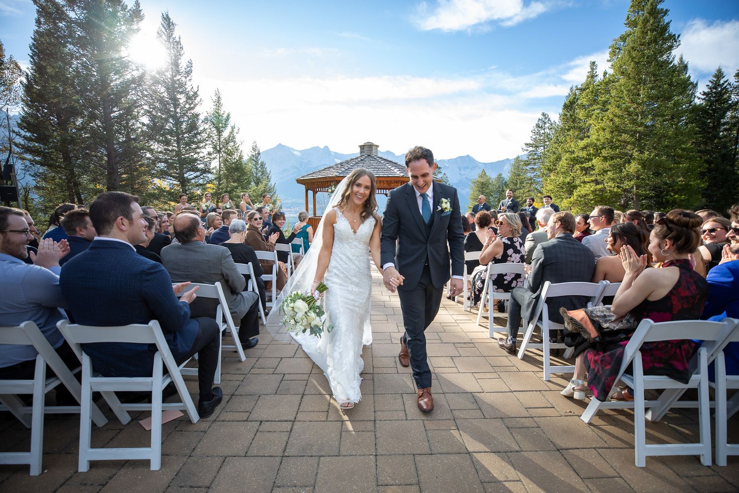 A joyful couple walks down an outdoor aisle at the Silvertip gazebo wedding ceremony with guests seated on white chairs. Canmore, AB.