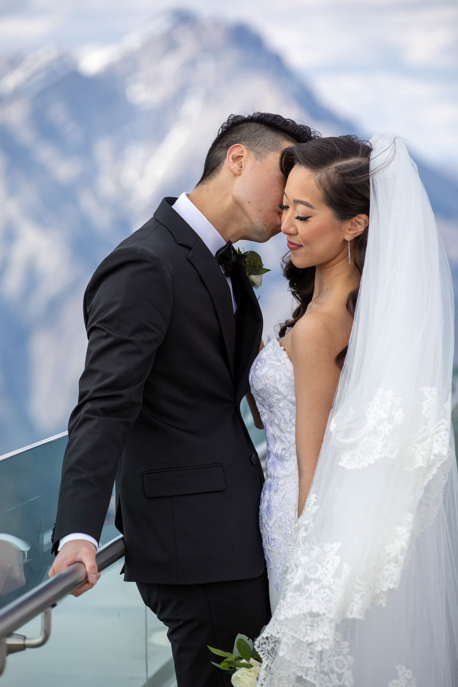 Groom in black suit kissing bride in lace wedding dress and veil on mountain balcony with snowy peaks backdrop.