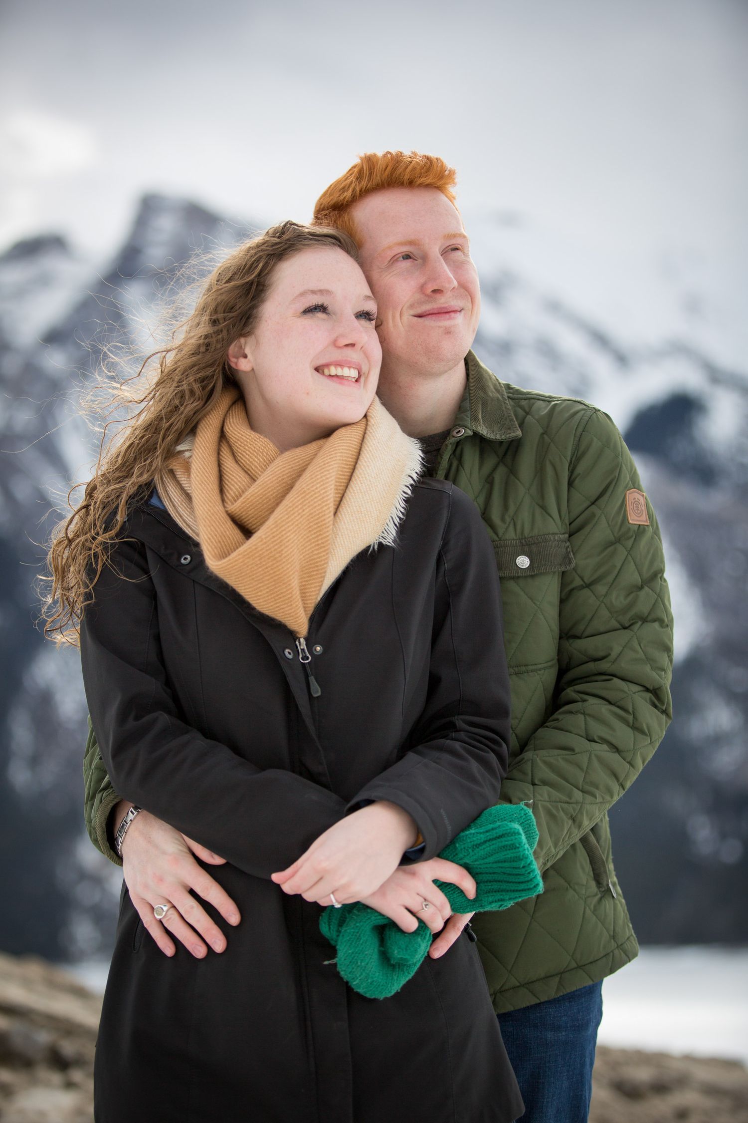 A happy couple embraces in front of snowy mountains, woman in black coat with yellow scarf, man in green jacket at Lake Minnewanka in Banff.