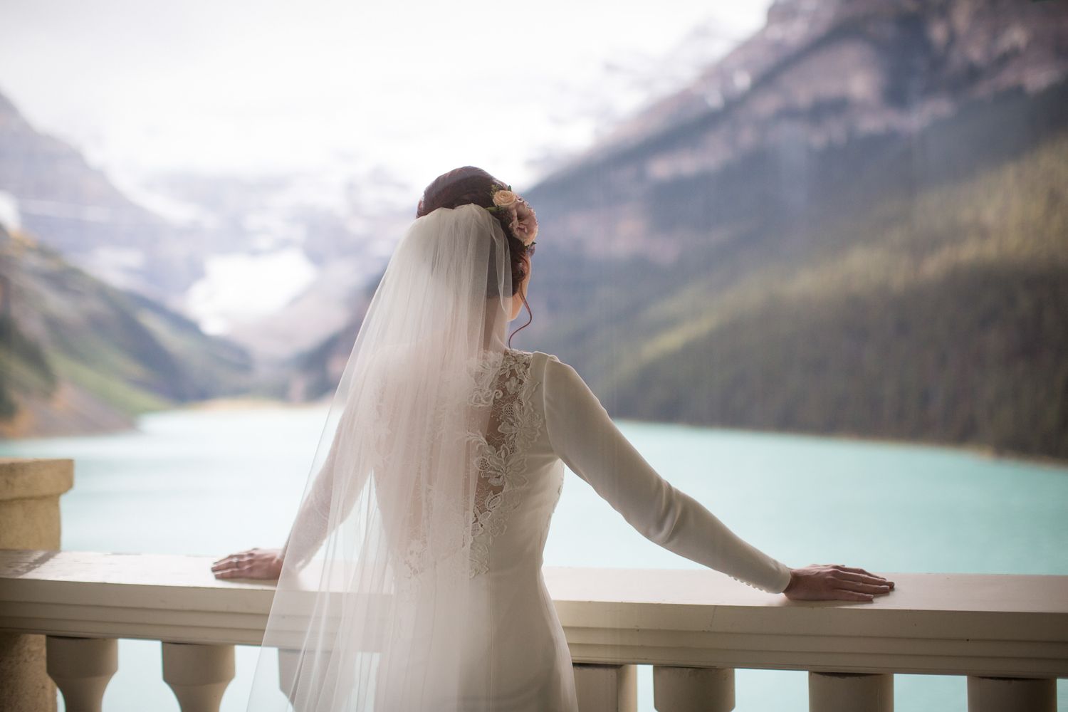 A bride in a white gown and veil stands on a deck overlooking a turquoise mountain lake in a scenic landscape. Chateau Lake Louise, AB