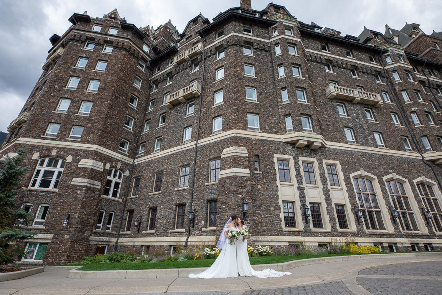 A couple in wedding attire stands outside a grand historic Fairmont Banff Springs Hotel in the background.