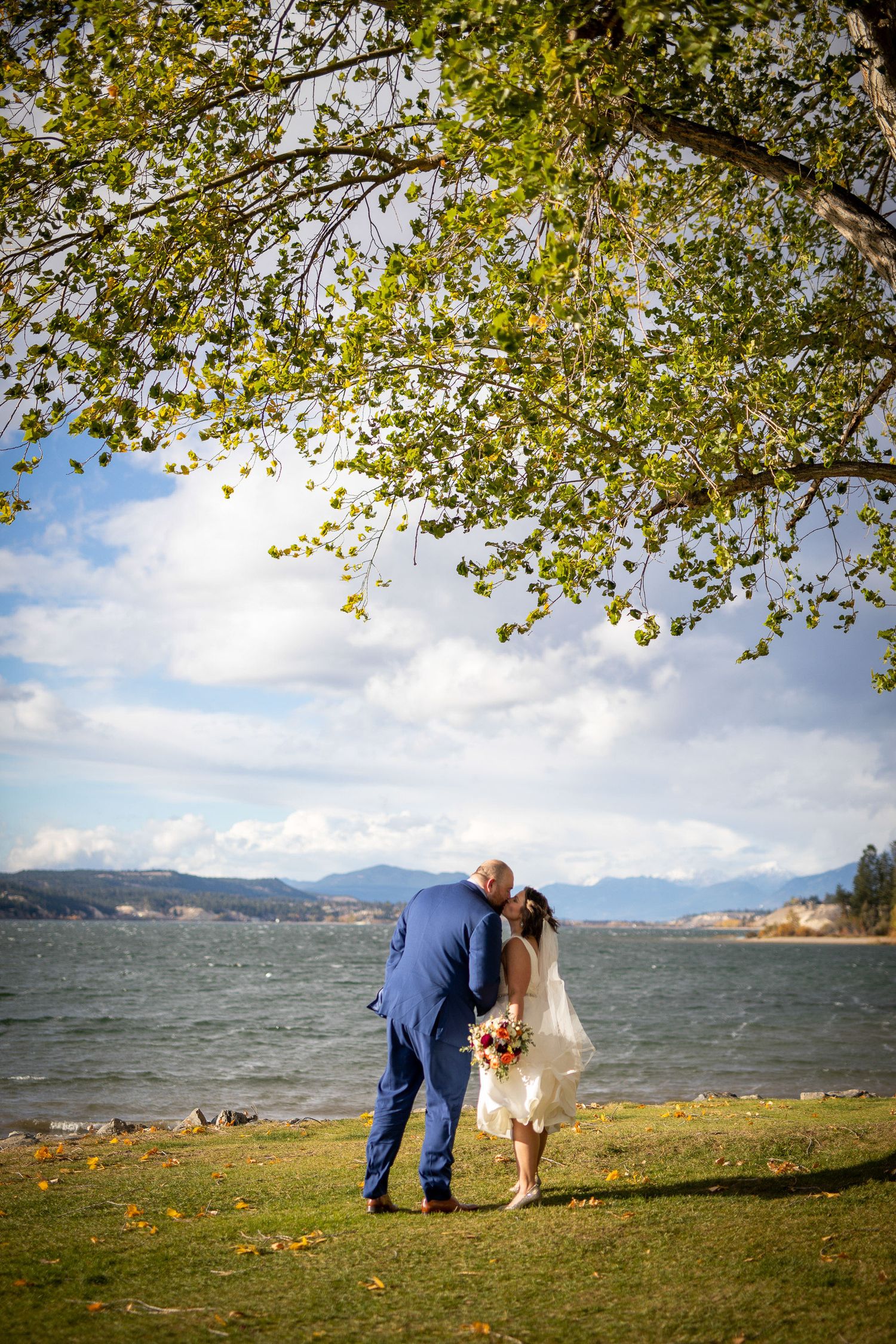 Couple in wedding attire bends down by a scenic mountain Windermere lake shoreline with autumn foliage and cloudy skies. Windermere, BC.
