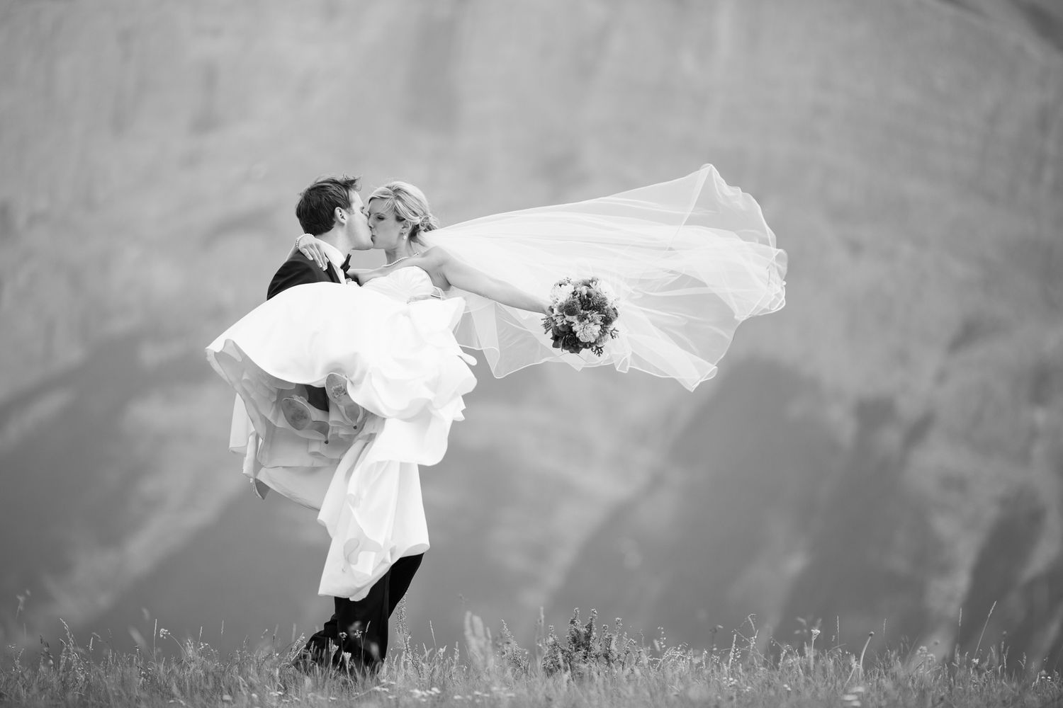 A romantic black and white wedding photo shows a couple embracing on a hillside as the bride's veil flows in the wind in Banff, alberta