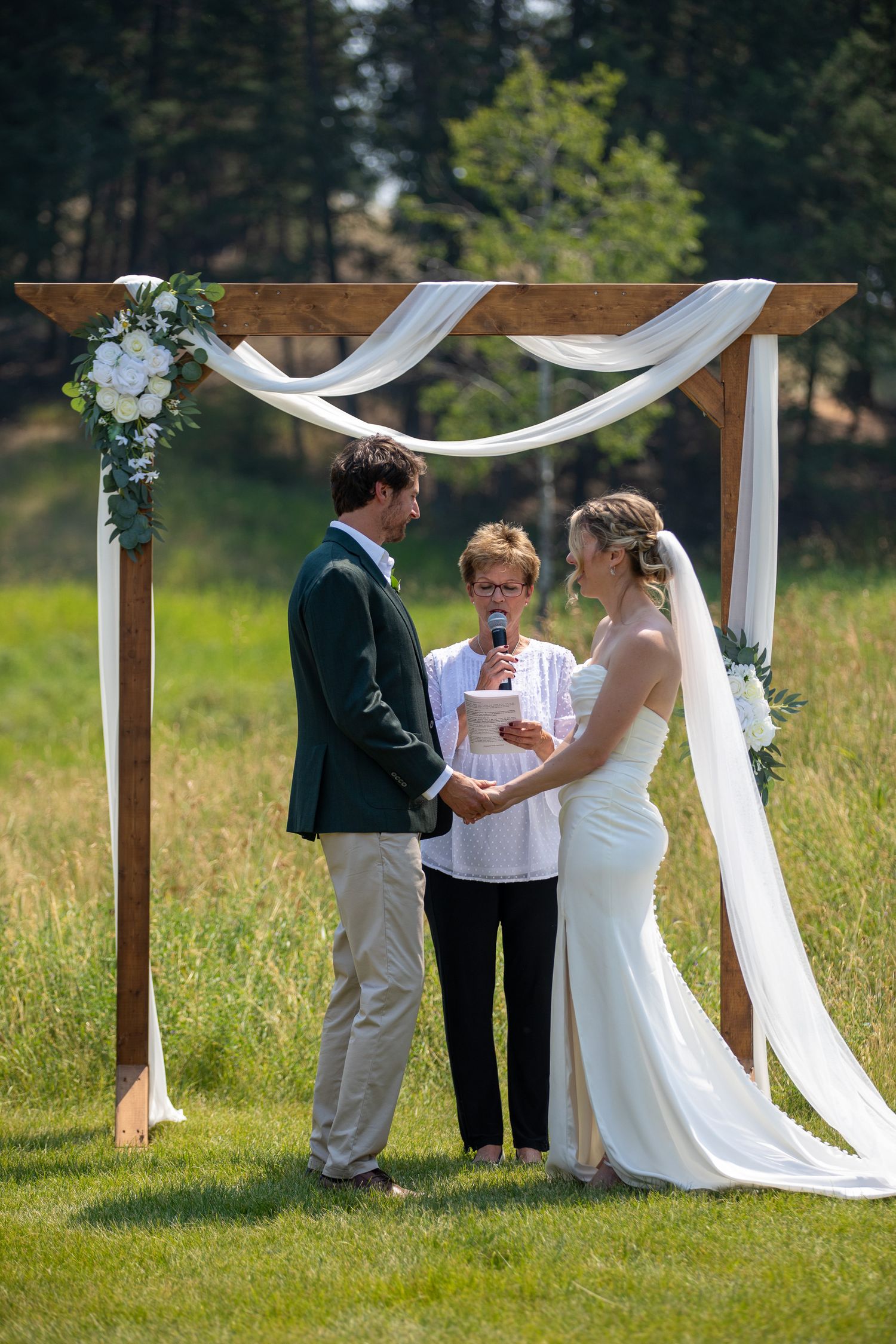 Outdoor wedding ceremony with bride and groom holding hands under a wooden arch with white draping and flowers at Swansea Ranch in Invermere