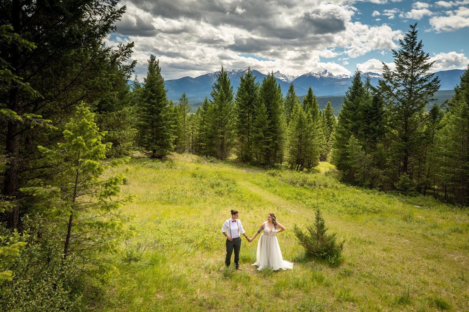 A couple walks through a scenic mountain meadow surrounded by evergreen trees and dramatic cloudy skies. Radium retreat, Radium, BC