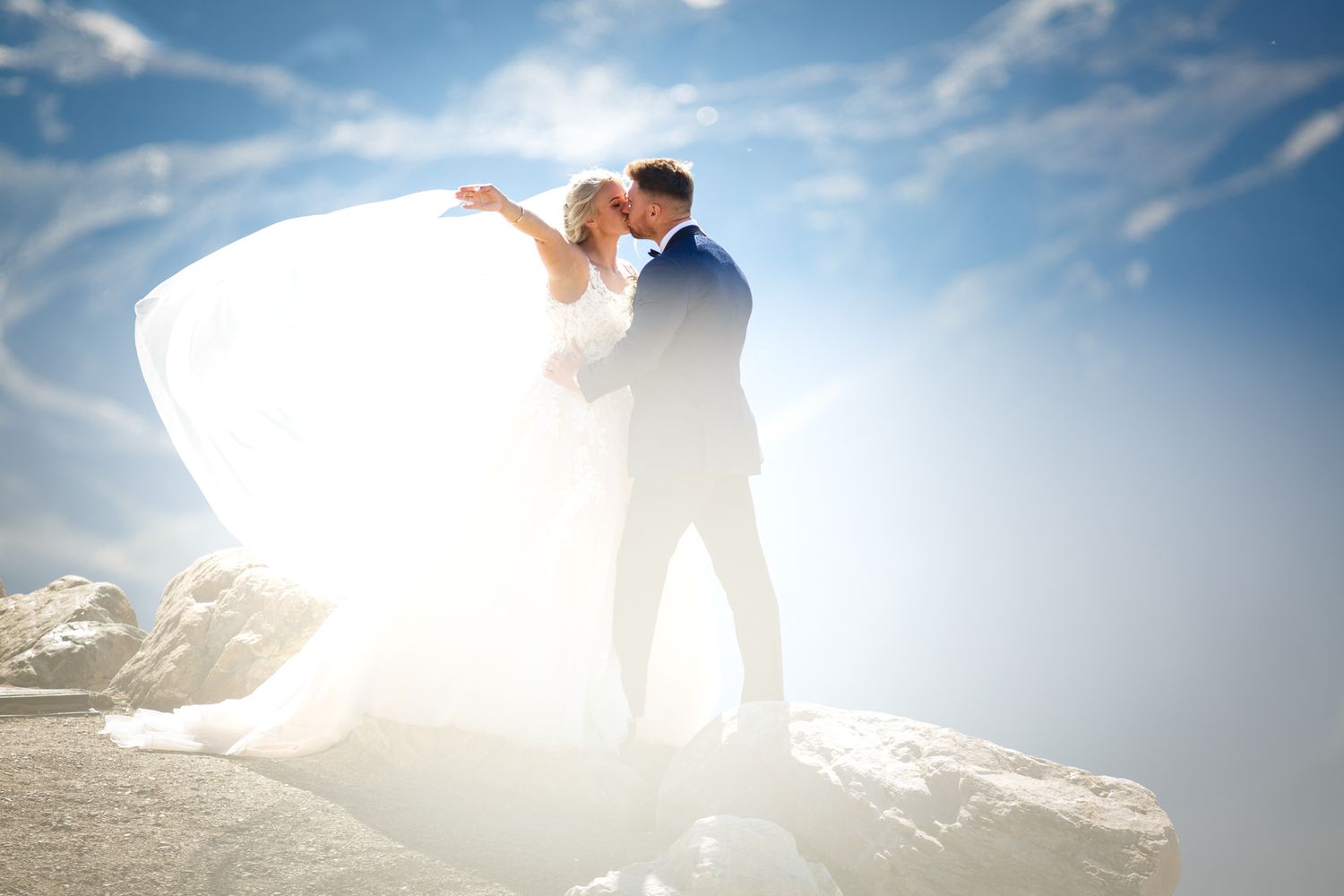 A couple in wedding attire shares a romantic silhouetted embrace against a bright blue sky and sunlight at Eagle Eye Resort in Golden, BC