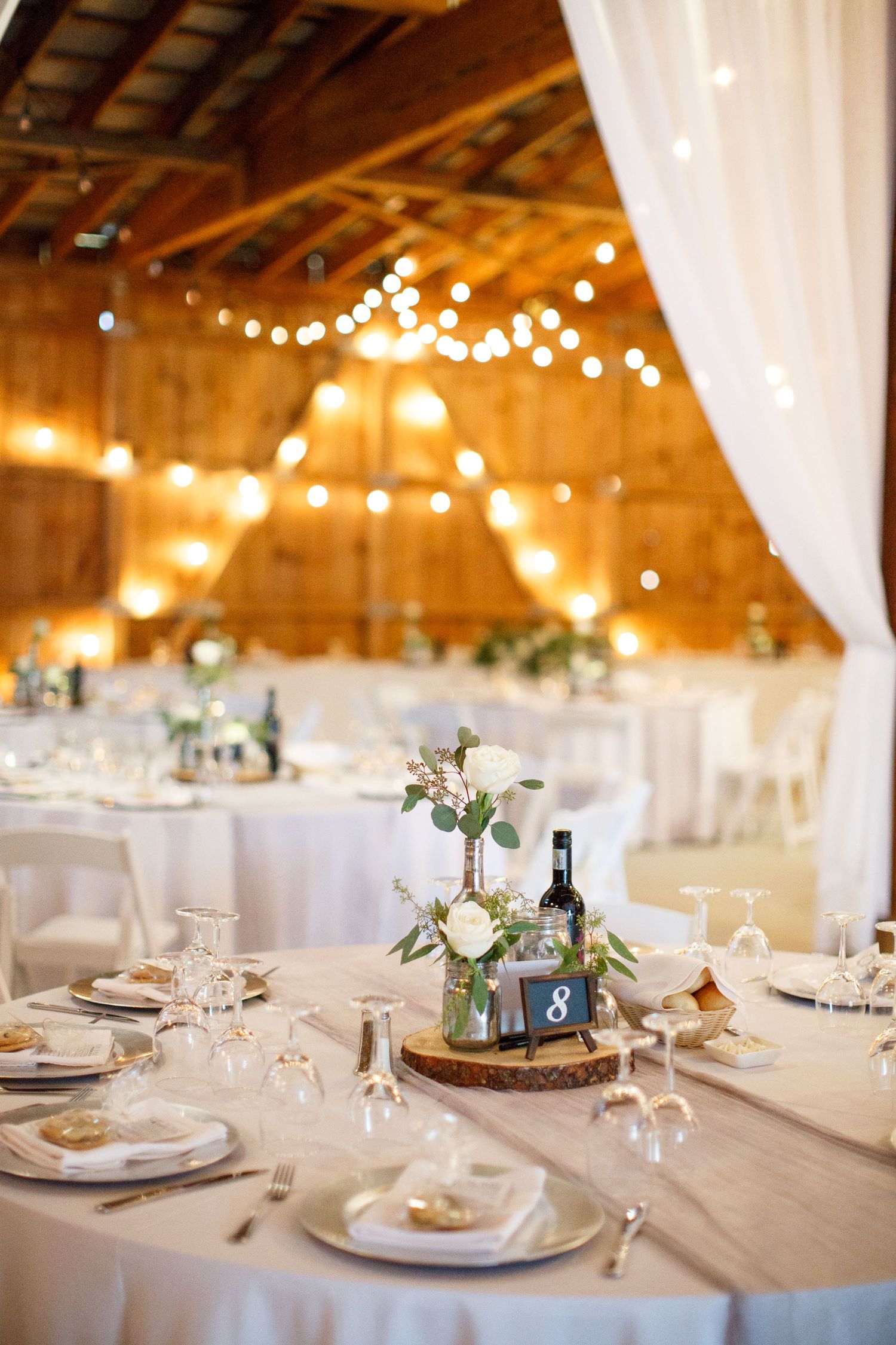 Rustic barn wedding reception with string lights, white draped fabric, floral centerpieces, and elegantly set round tables.