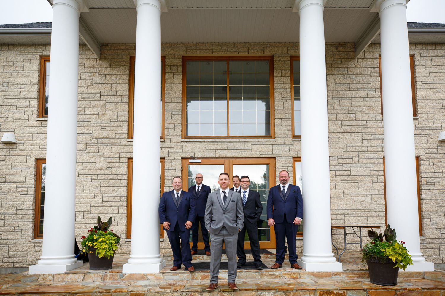 Groom in gray suit stands with groomsmen in navy suits on steps of elegant stone venue with white columns at Cherry creek estate