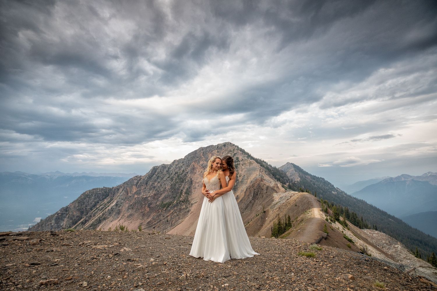 Two brides in white gowns embrace on a rocky mountain summit with dramatic cloudy skies and peaks behind them at Kicking Horse Resort, BC