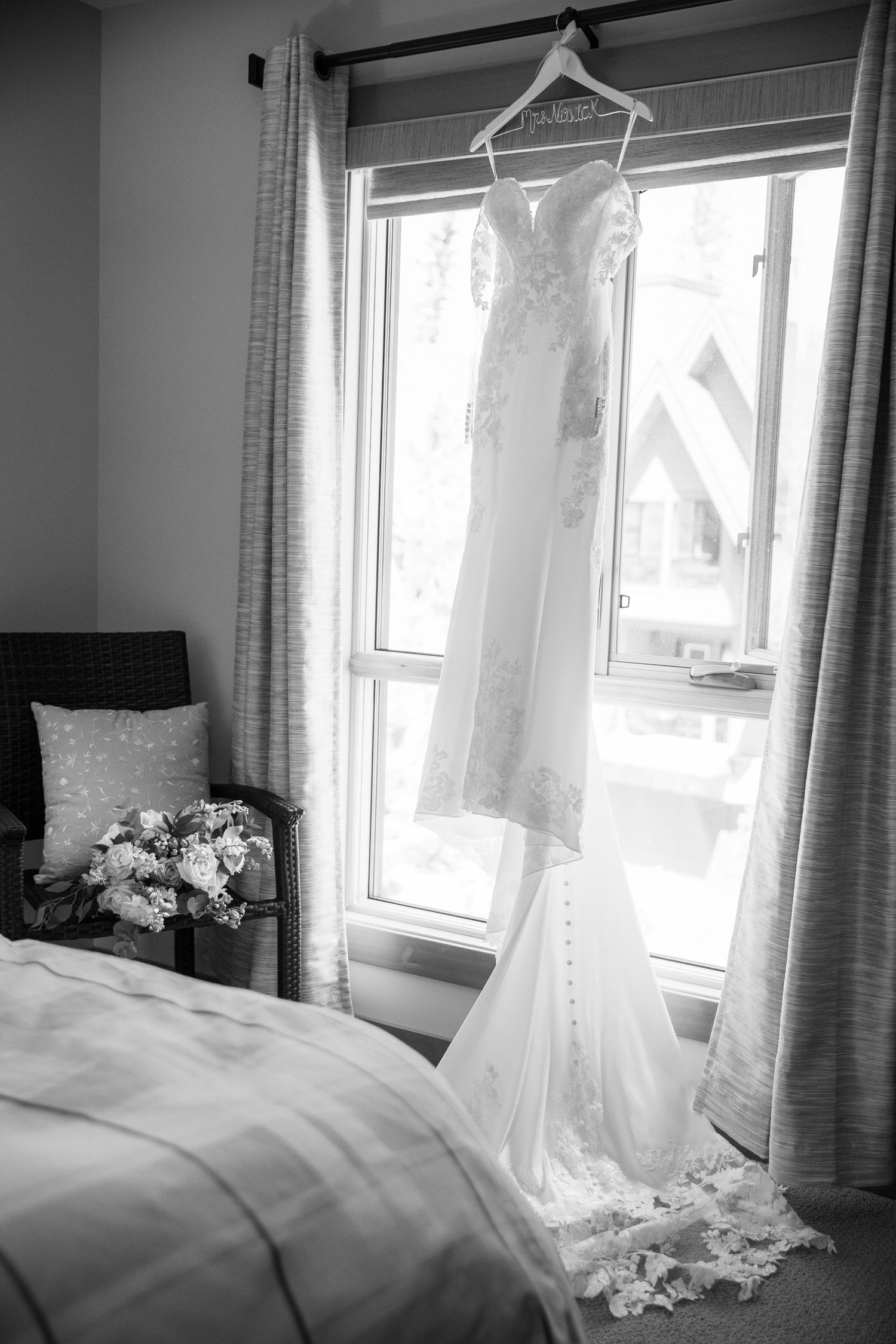 Elegant lace wedding dress hanging by a window in black and white, with a bridal bouquet on a chair beside a bed In Canmore