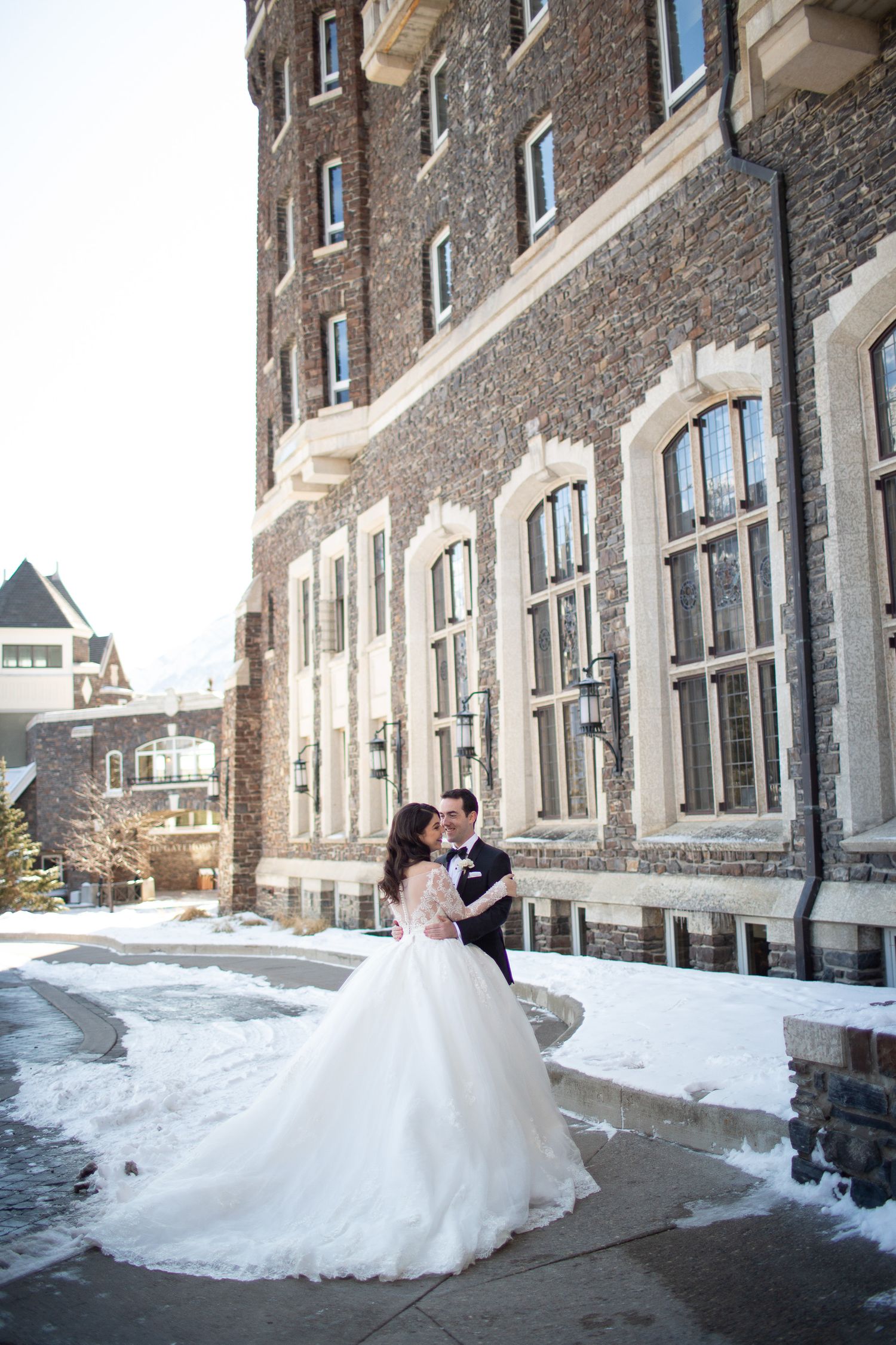 A couple shares a romantic moment outside a historic brick building on a snowy winter day. Fairmont Banff Springs Hotel in Banff.