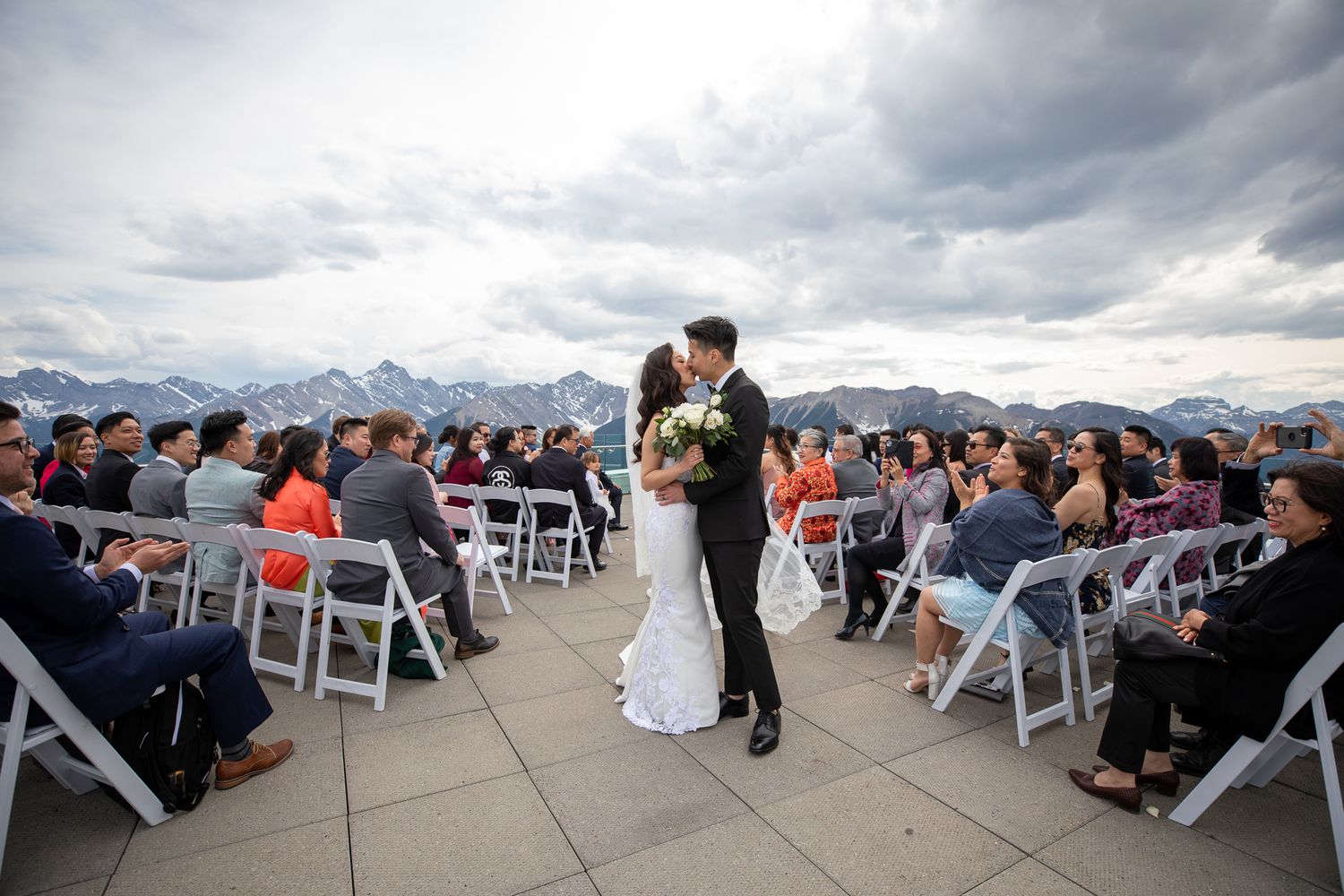 Couple shares first kiss at outdoor rooftop wedding ceremony with mountain backdrop and seated guests under cloudy skies.