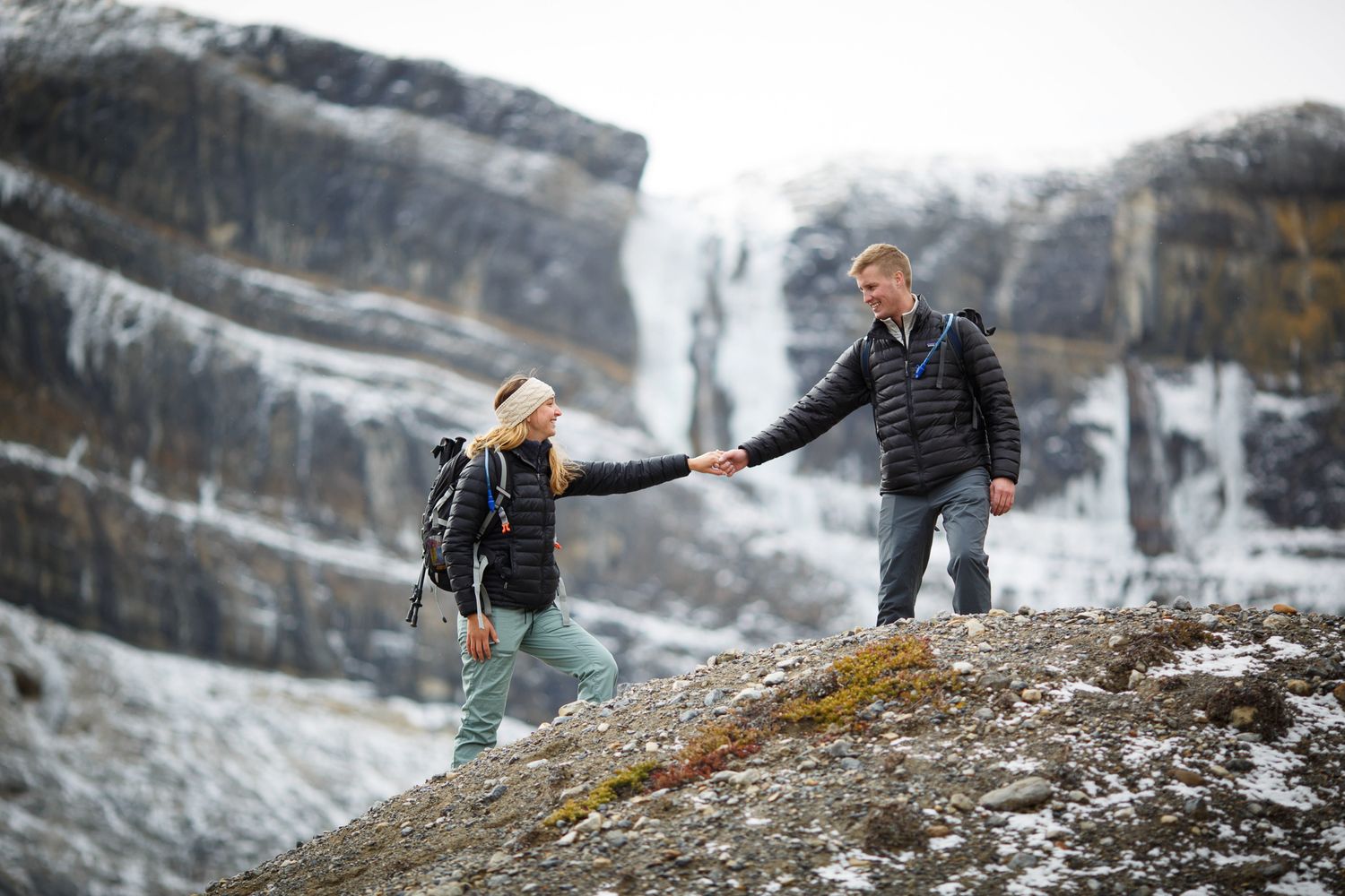 Two people hold hands while hiking on rocky terrain with a snowy mountain waterfall in the background at Bow Lake in Banff National Park.