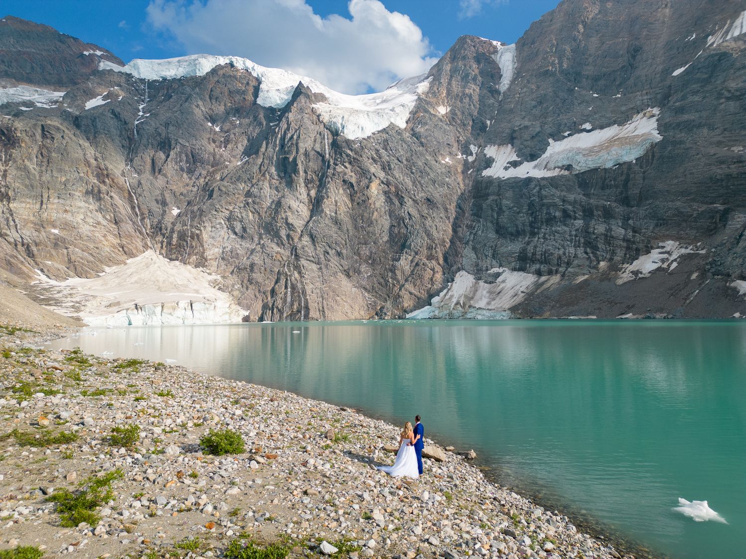 Couple in wedding attire standing beside a stunning turquoise glacial lake surrounded by rocky mountains and snow-capped peaks. Panorama, BC