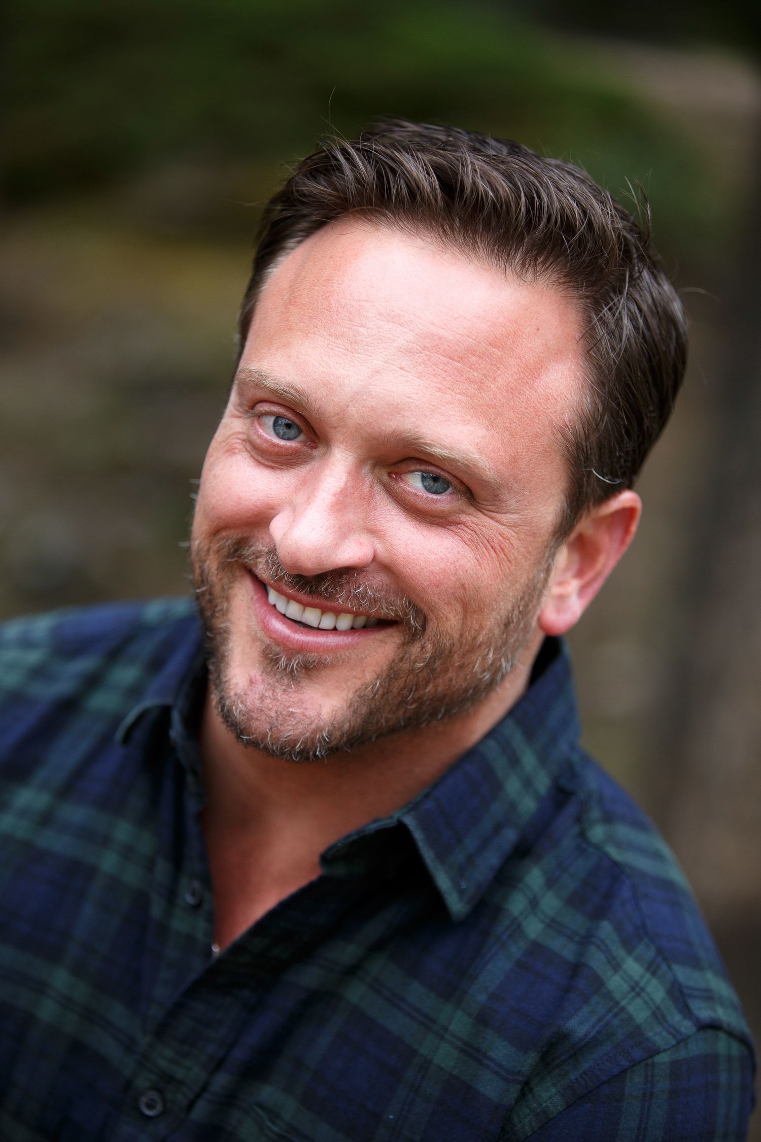 Smiling man with dark hair and beard wearing a blue plaid shirt outdoors.
