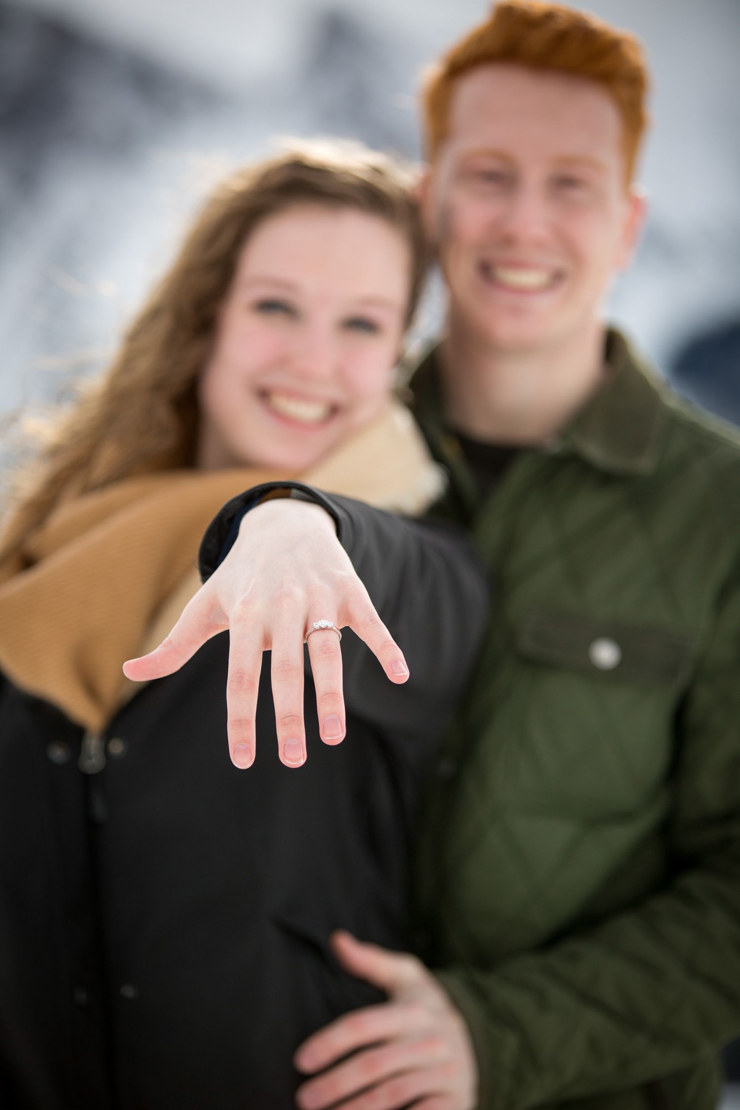 Happy couple showing off engagement ring in snowy winter setting, woman displaying diamond ring on her finger at Lake Minnewanka in Banff.