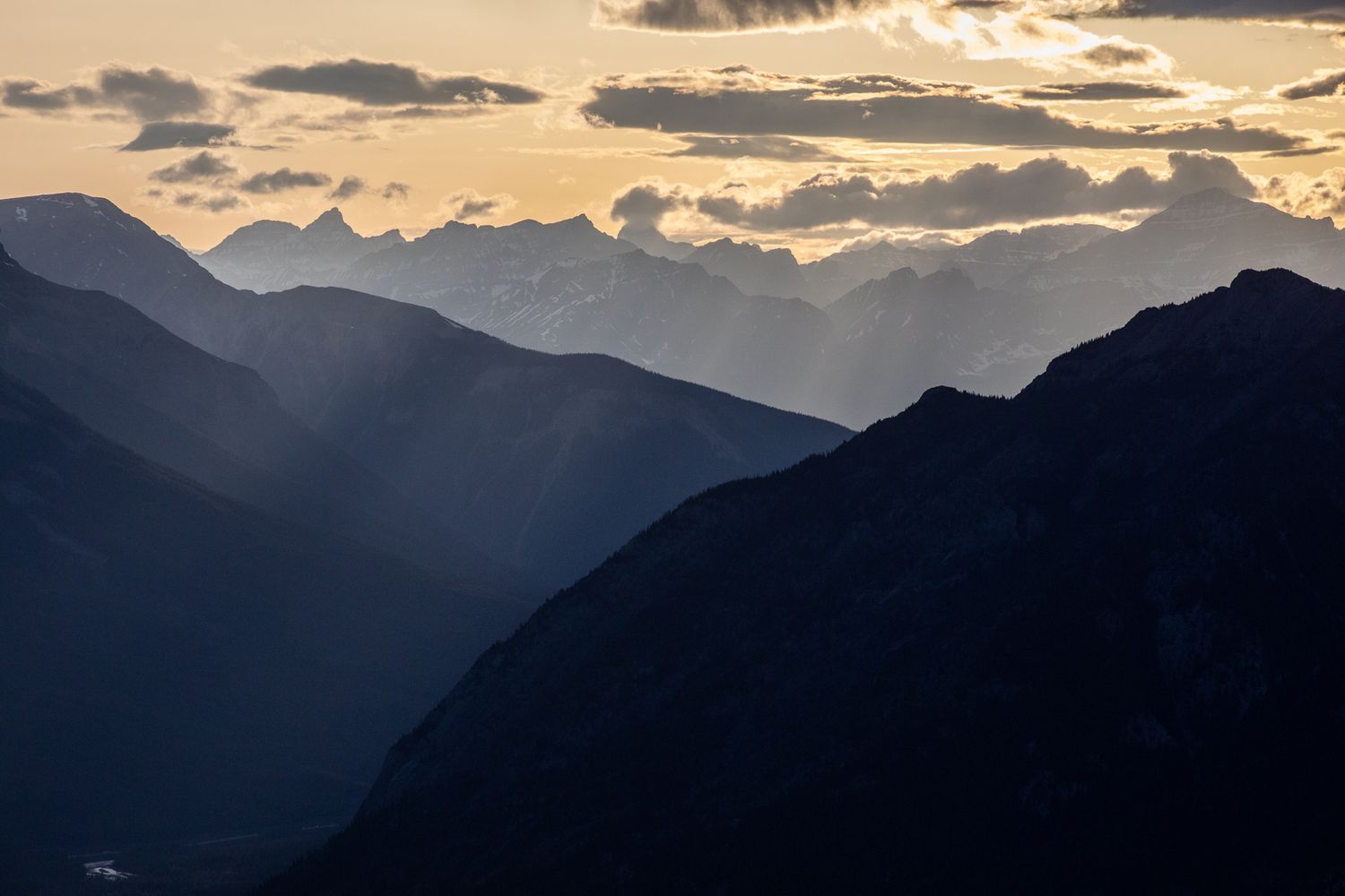 Dramatic mountain sunset with golden rays piercing through clouds over silhouetted alpine peaks and layered valleys.