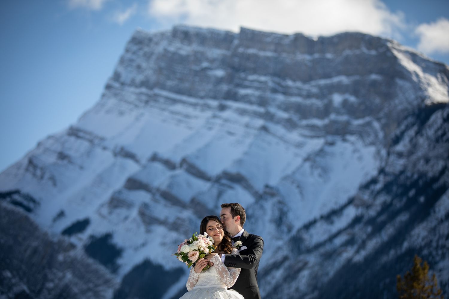 A couple embracing in front of a snow-capped mountain with a cloudy sky in the background at Tunnel reservoir in Banff.