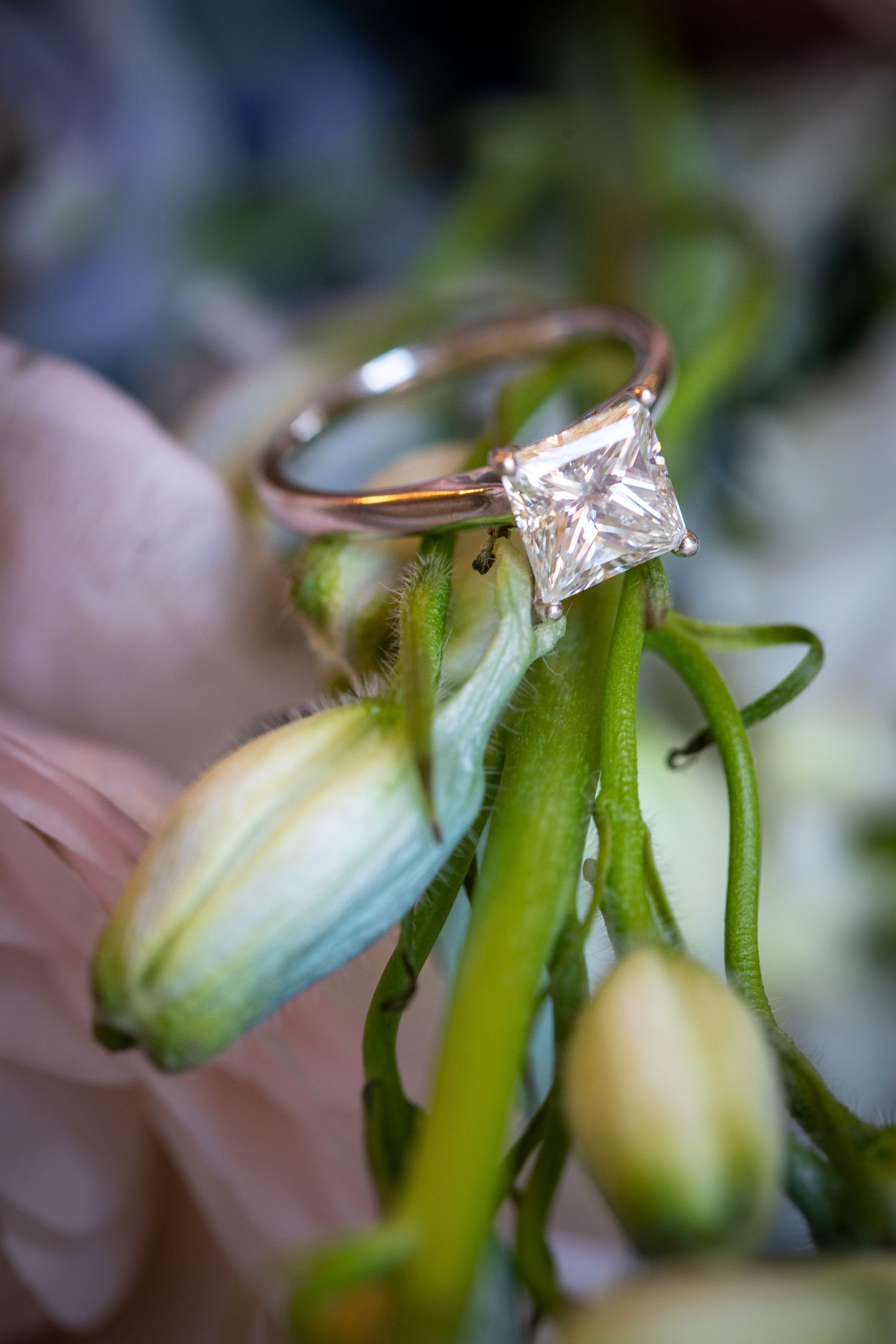 Princess cut diamond solitaire engagement ring resting on green flower buds in a soft floral arrangement.