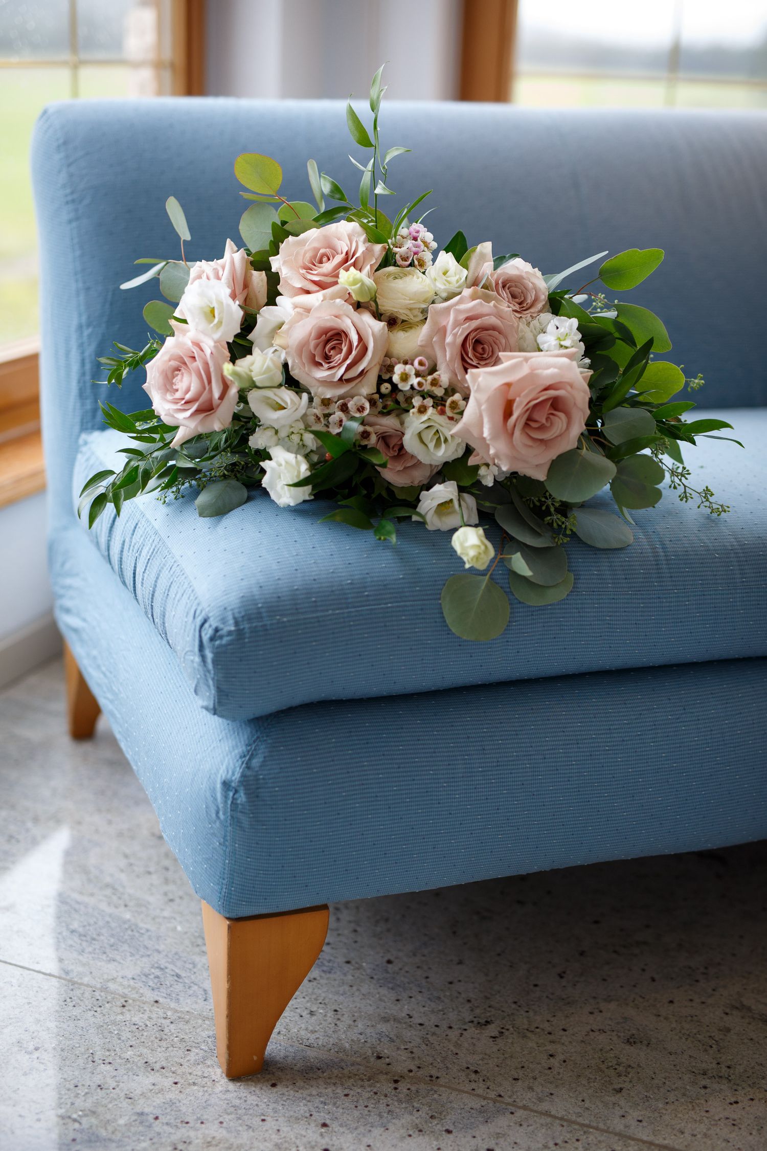 Elegant bridal bouquet of blush pink roses and white flowers on a light blue sofa near a sunlit window.