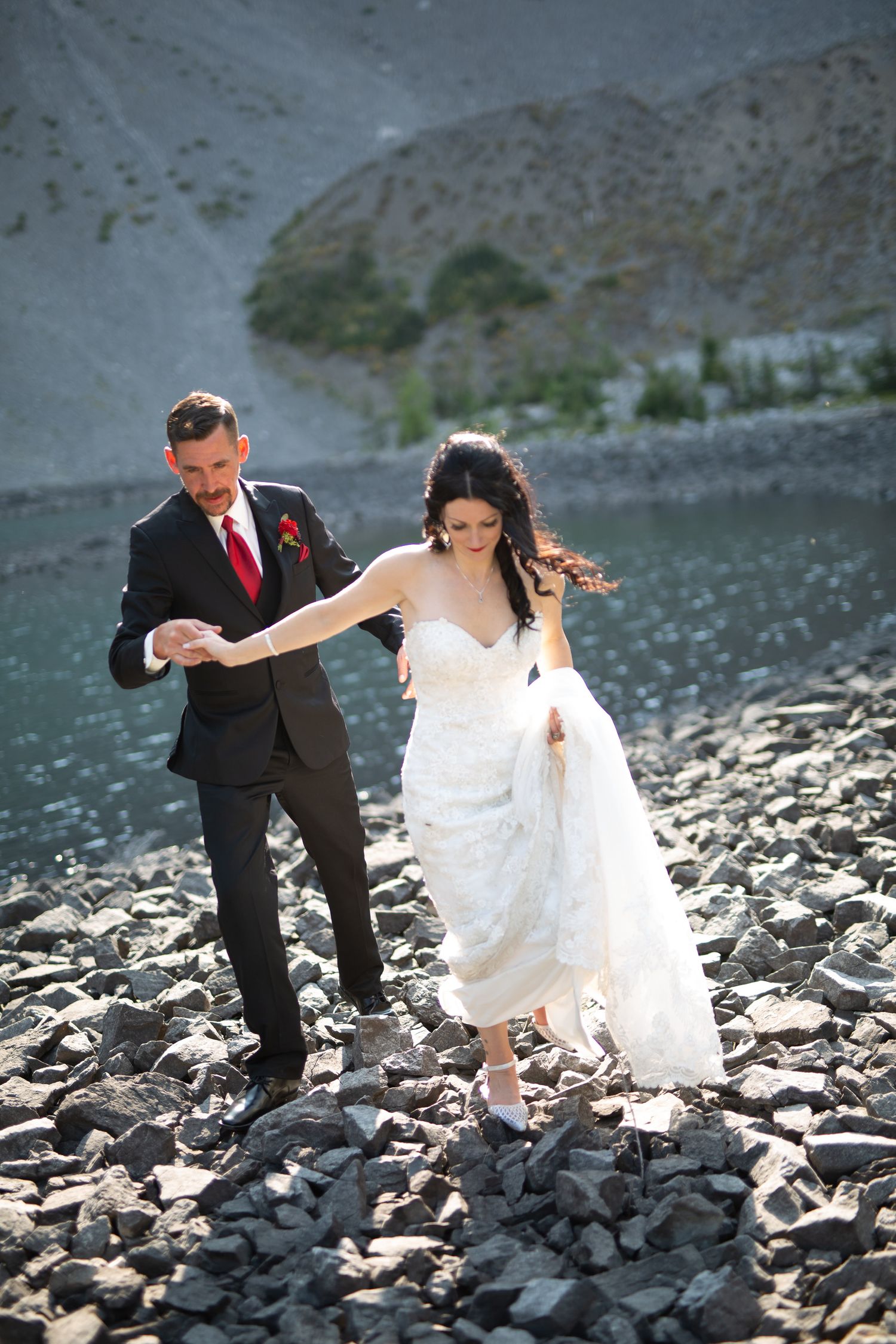 Bride and groom walking on rocky shoreline near mountain lake, groom in black suit with red tie holding bride's handin Kananaskis, Alberta.