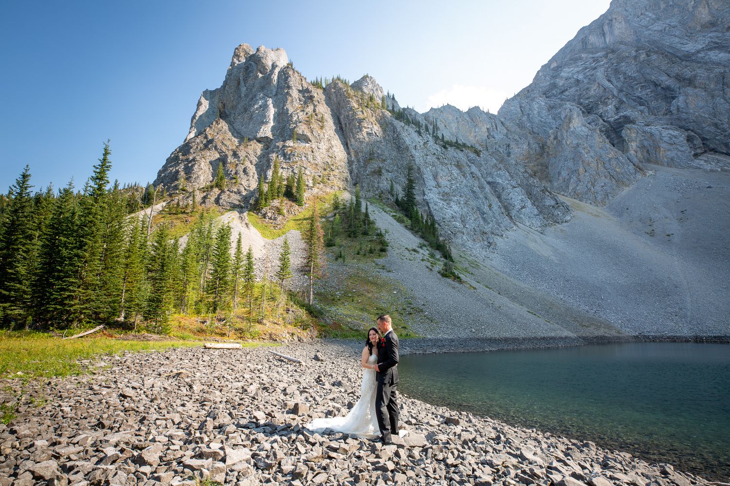 Bride and groom embrace by a rocky alpine lake with dramatic mountain peaks and evergreen trees in the background in Kananaskis, Alberta.