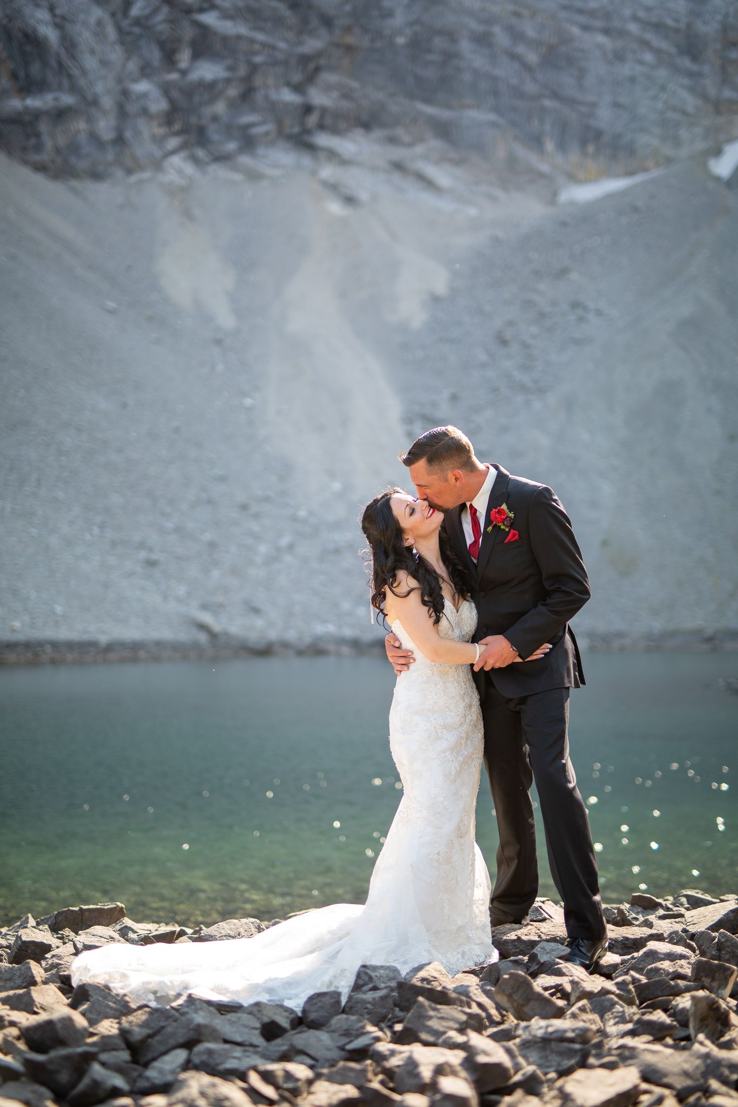 Bride and groom share a romantic kiss by a turquoise mountain lake, surrounded by rocky terrain and snowy cliffs in Kananaskis, Alberta.