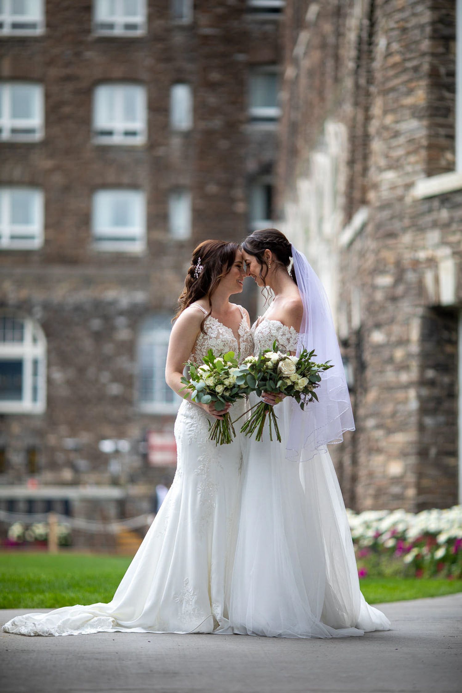 Two LGBTQ brides in white wedding dresses standing together holding bouquets of flowers on stone steps at the Fairmont Banff Springs Hotel.