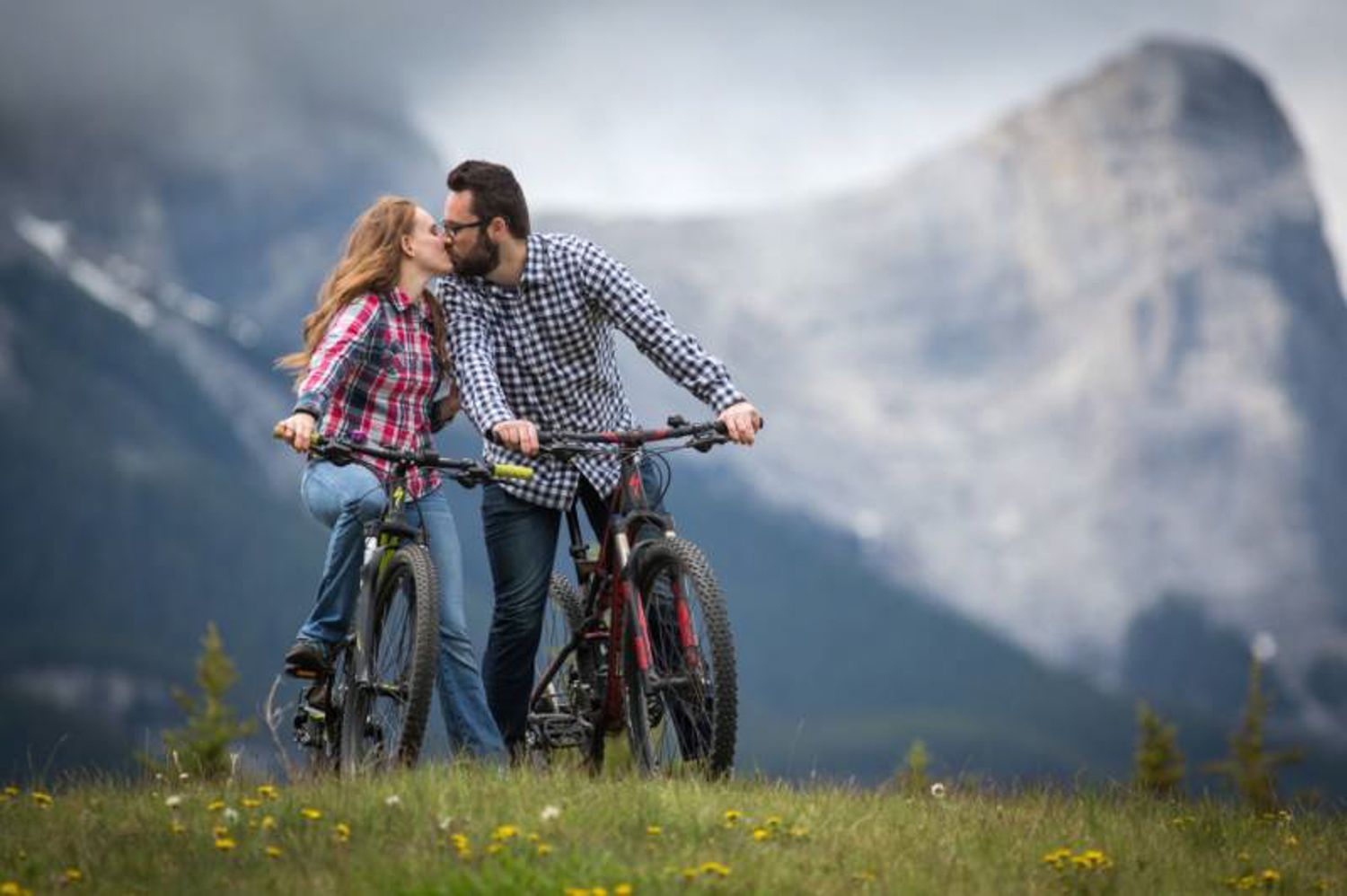 Couple sharing a romantic moment with bicycles in a scenic mountain meadow with wildflowers and dramatic clouds in Canmore Quarry Lake Park.