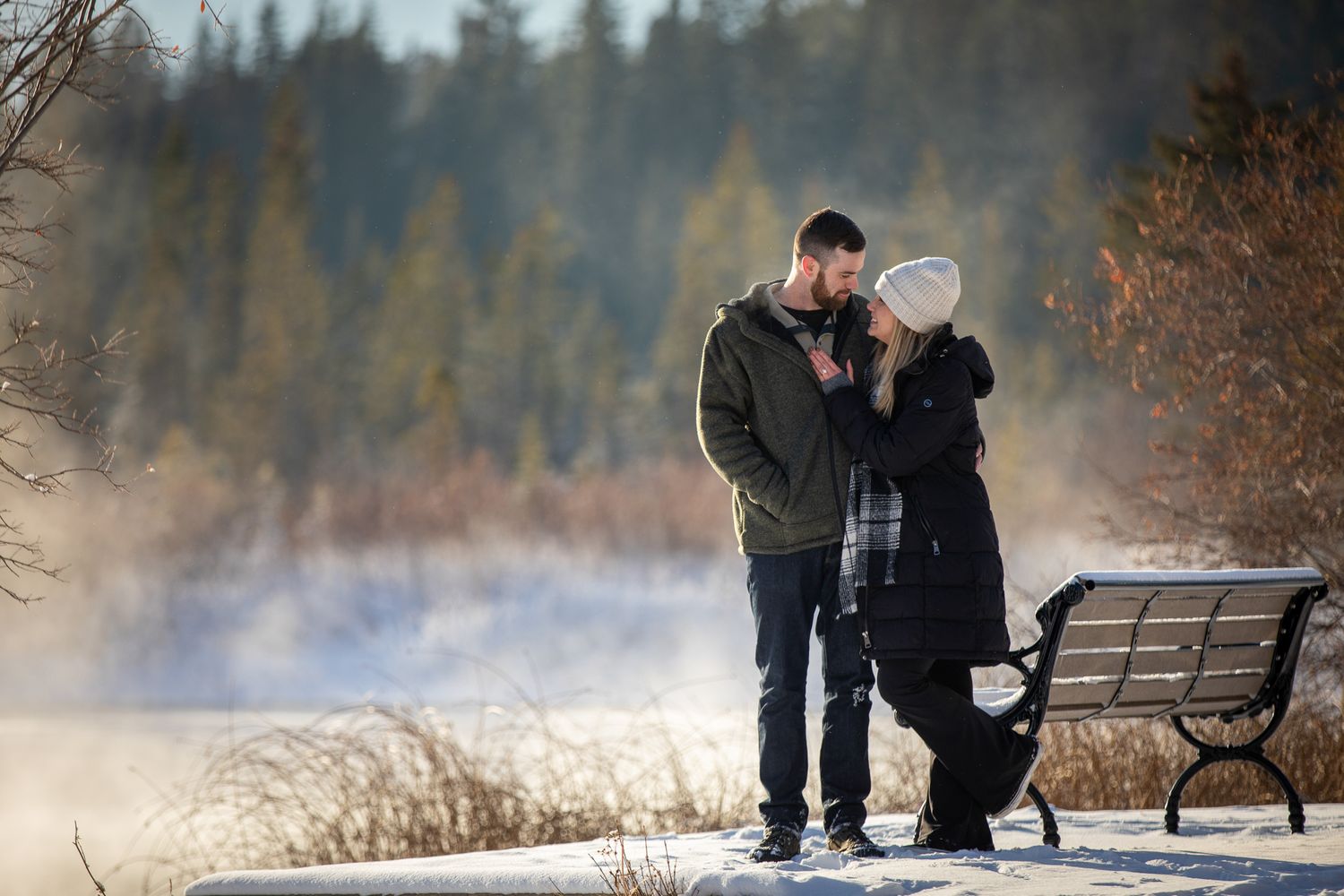 A couple stand along a snowy lakeshore of Vermilion lake watching a geyser erupt in the misty winter landscape in Banff.