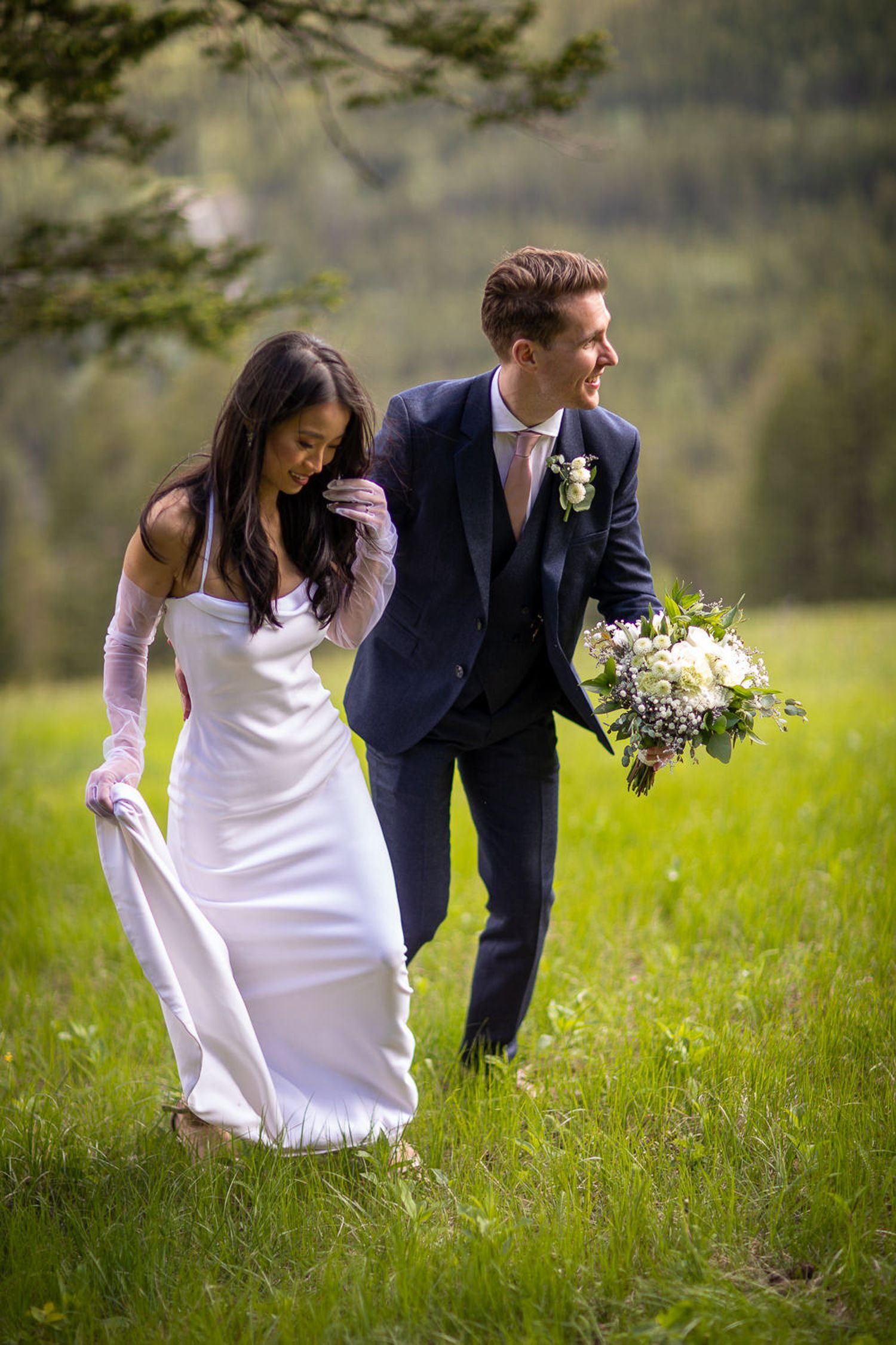 A bride and groom share a playful moment together in a lush green meadow during their outdoor wedding photoshoot. Mount Norquay lookout