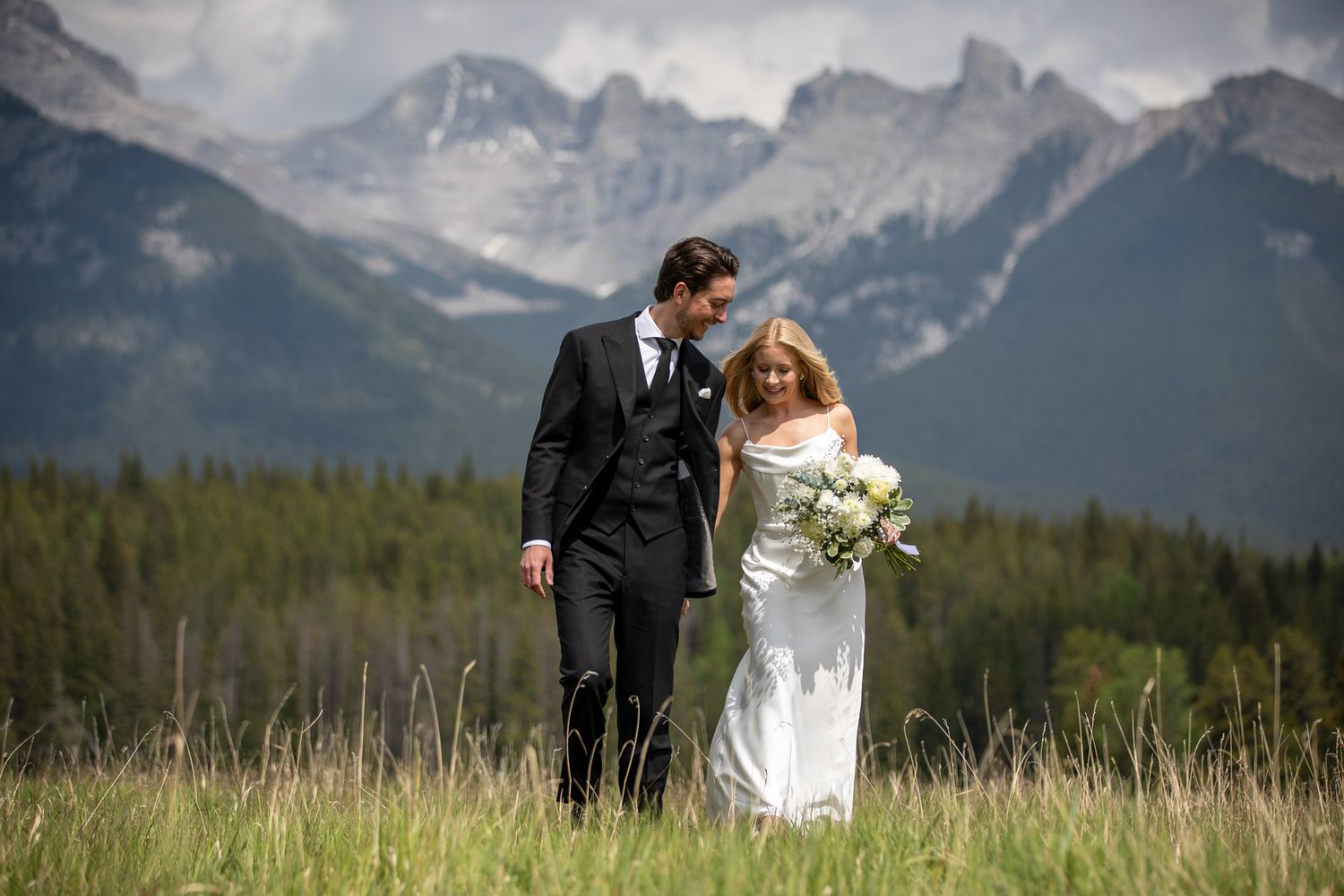 A couple in wedding attire walks through a scenic mountain meadow with dramatic Rundle peaks in the background. Banff National Park. AB.
