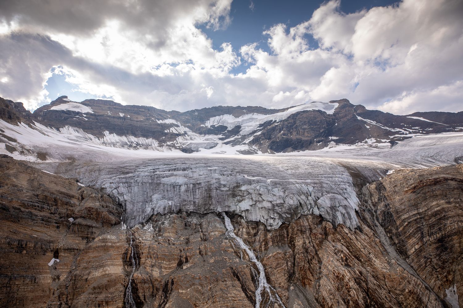 Dramatic glacier flowing over rocky cliffs in the Canadian Rockies, with snow-capped mountains and cloudy sky above. Lake-of-Hanging-Glacier