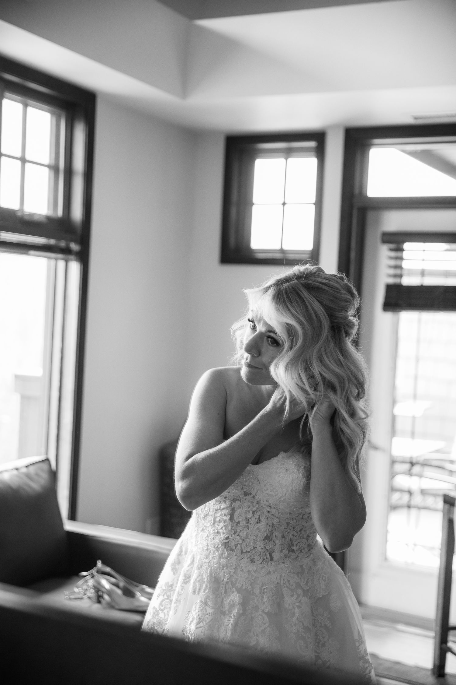 Black and white photo of a bride in a lace wedding gown adjusting her earring near sunlit windows. Copperpoint resort,  Invermere.