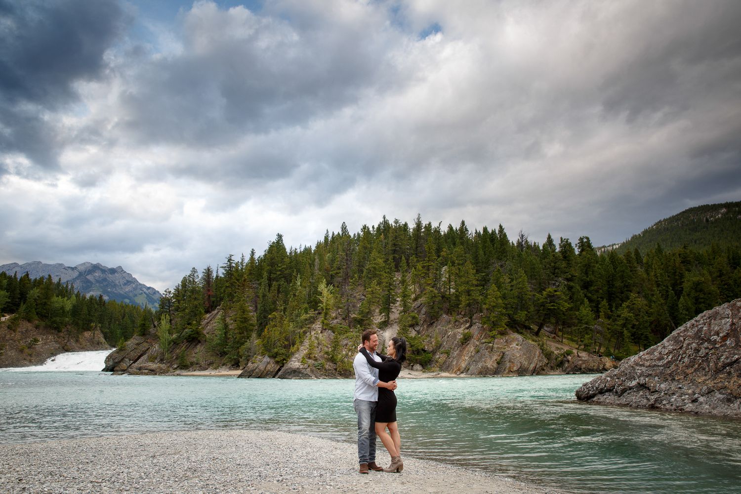 Couple embracing on a rocky riverbank with turquoise water, of Bow Falls in Banff and evergreen forest, and dramatic cloudy mountain sky.