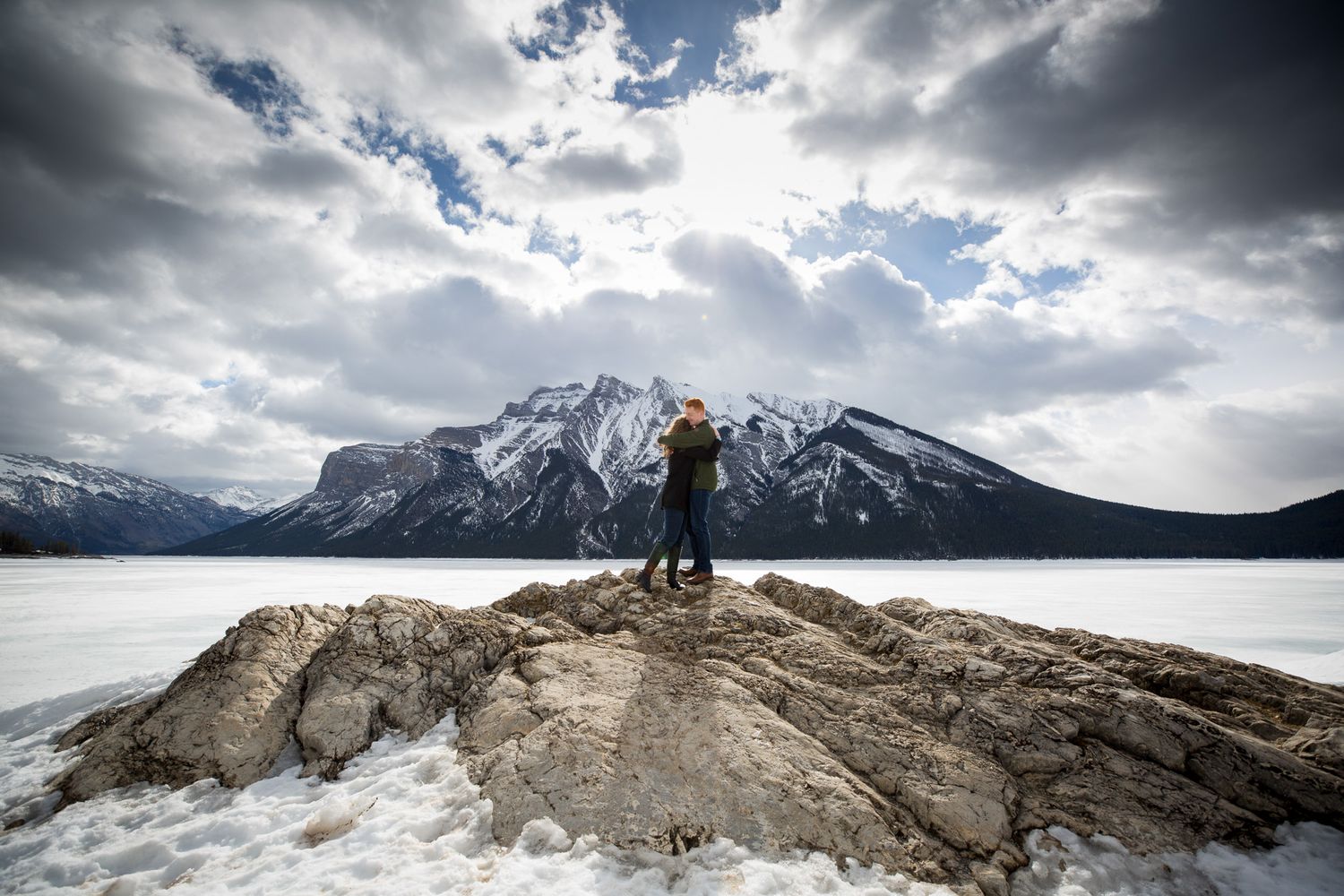 Person standing on rocky outcrop overlooking frozen lake and snow-capped mountains under dramatic cloudy sky at Lake Minnewanka in Banff.