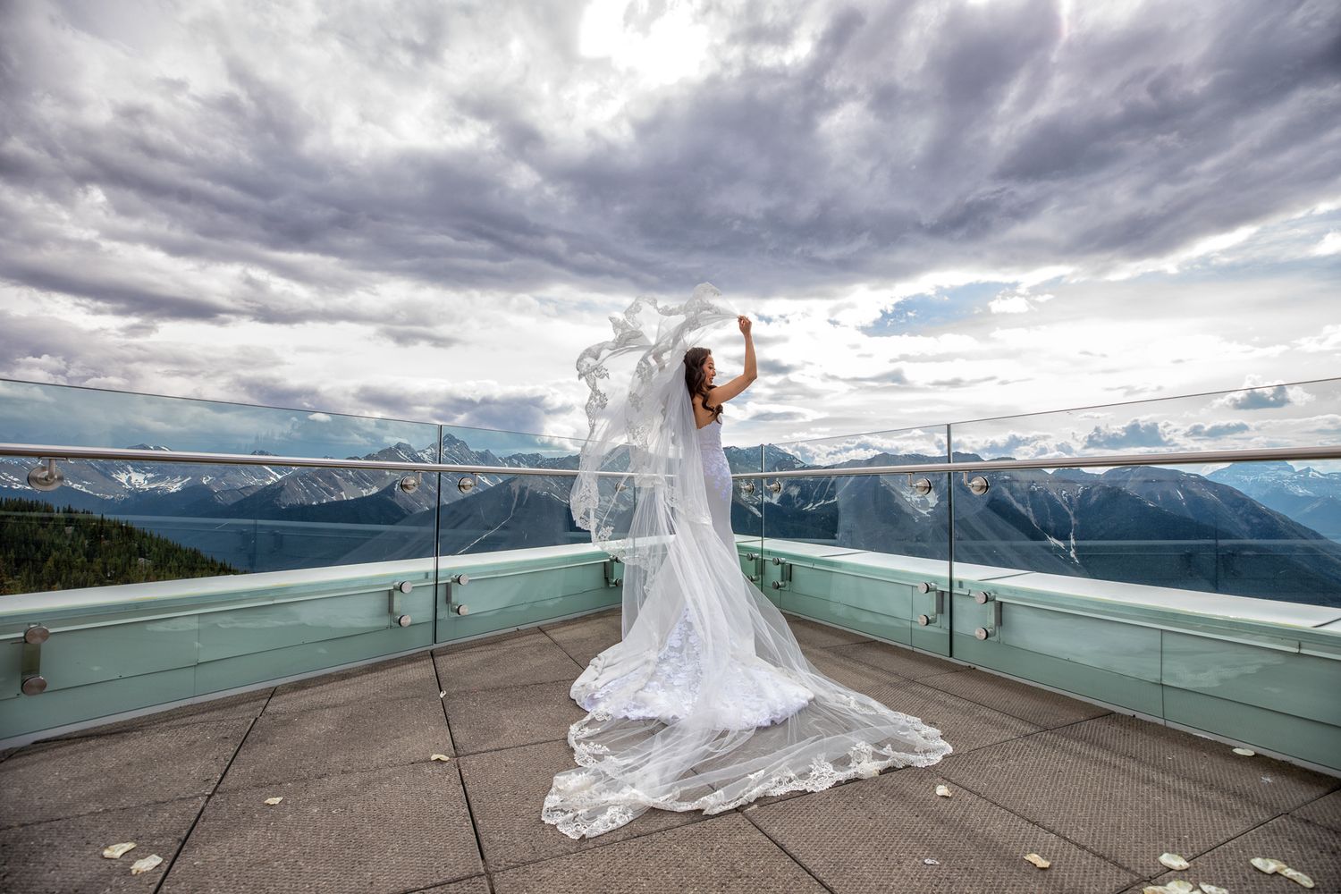 Bride in white gown and flowing veil stands on mountain overlook terrace with dramatic cloudy sky and alpine valley views.