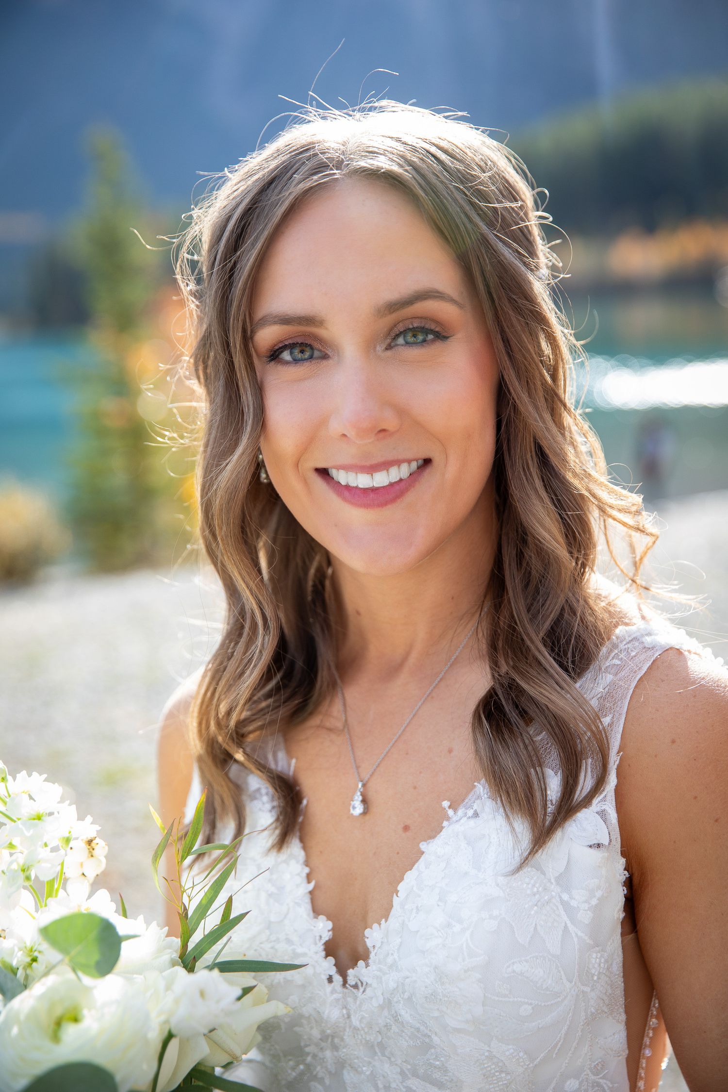 A bride in a white lace wedding dress holds a bouquet of white flowers against a scenic lakeside background at Quarry lake in Canmore, AB.