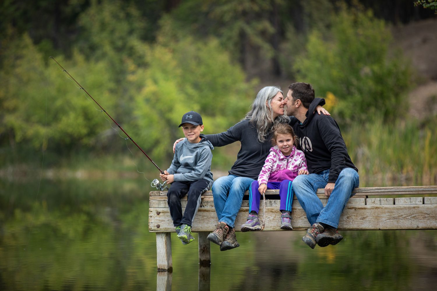 Family sitting together on a wooden dock by a tranquil pond surrounded by lush greenery at Lilian lake in Invermere, BC