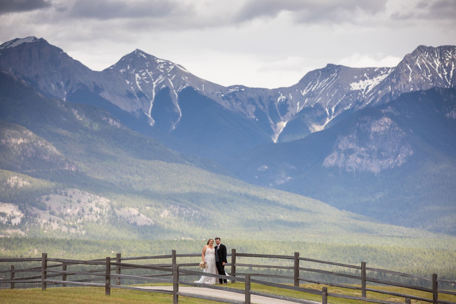A wedding couple stands on a road overlooking majestic snow-capped mountains in the distance. Brady Creek ranch. Invermere, BC.