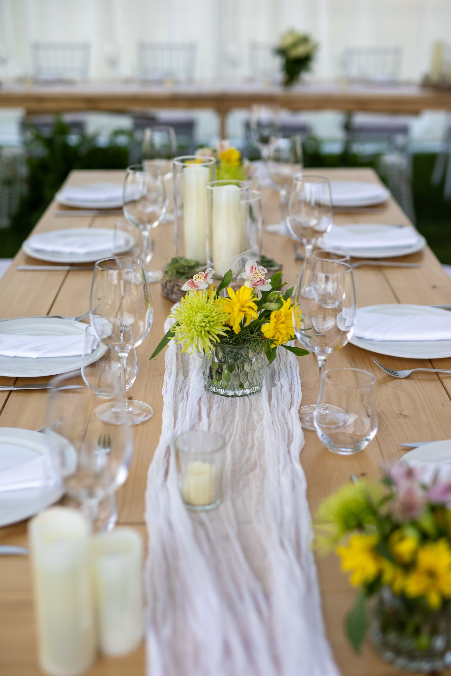 Elegant wooden wedding reception table set with white plates, wine glasses, candles, and yellow floral centerpieces on a white runner