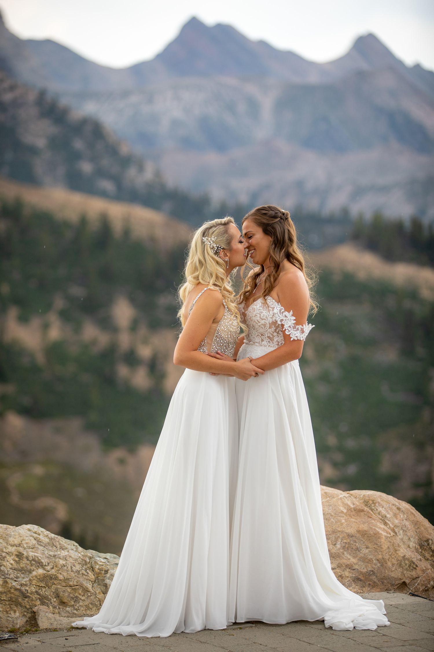 Two brides in white lace gowns share a romantic kiss with stunning mountain scenery backdrop at Kicking Horse Mountain Resort, Golden, BC
