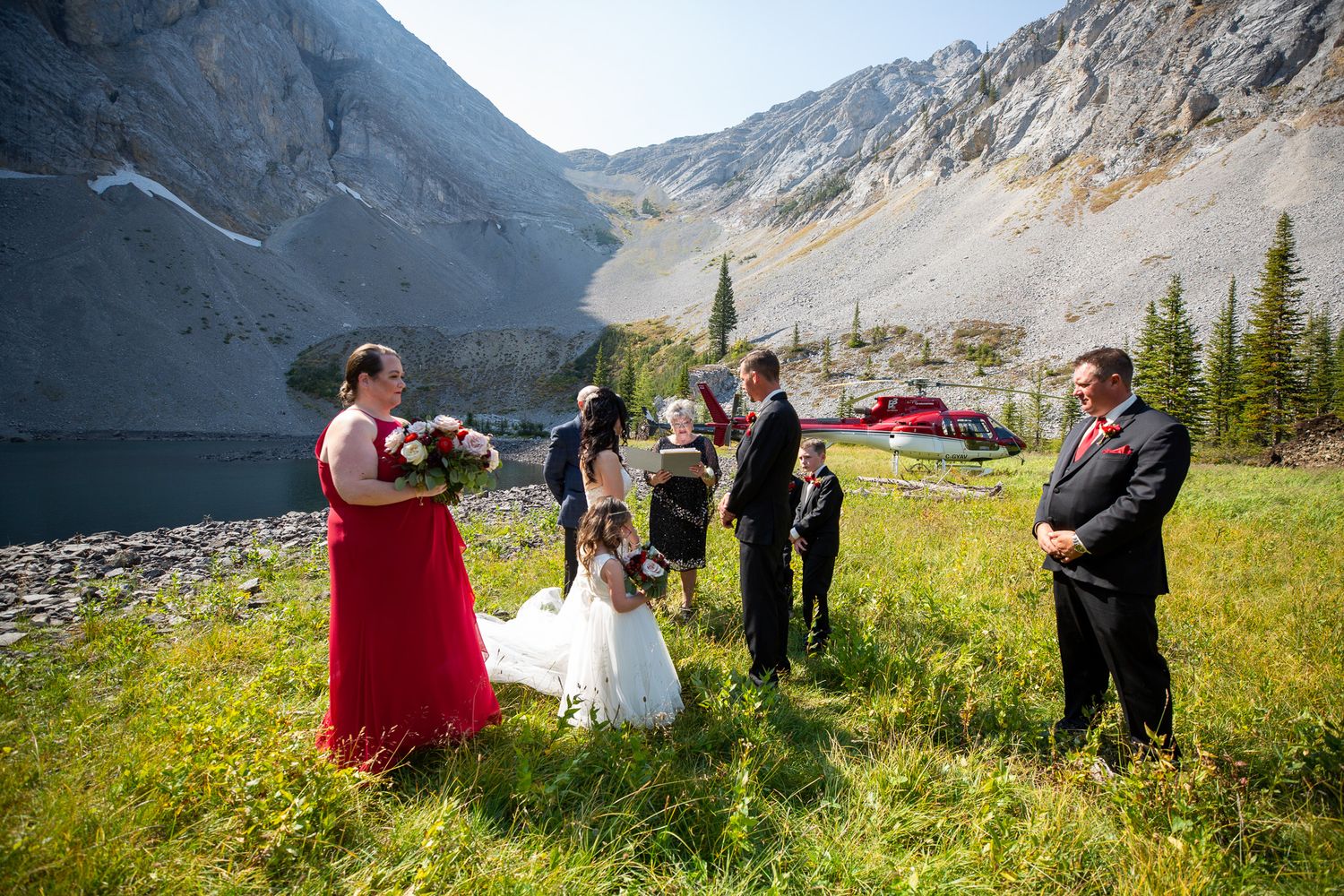 Outdoor mountain wedding ceremony in a green meadow with bridal party in red and black attire, helicopter in background in Kananaskis, AB