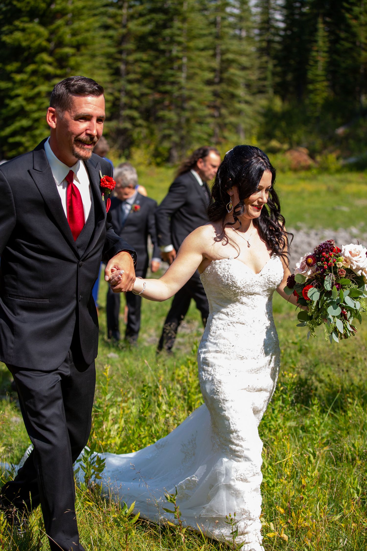 Bride and groom hold hands walking through a mountain meadow, bride in lace gown holding red bouquet, groom in black suit in Kananaskis, AB.