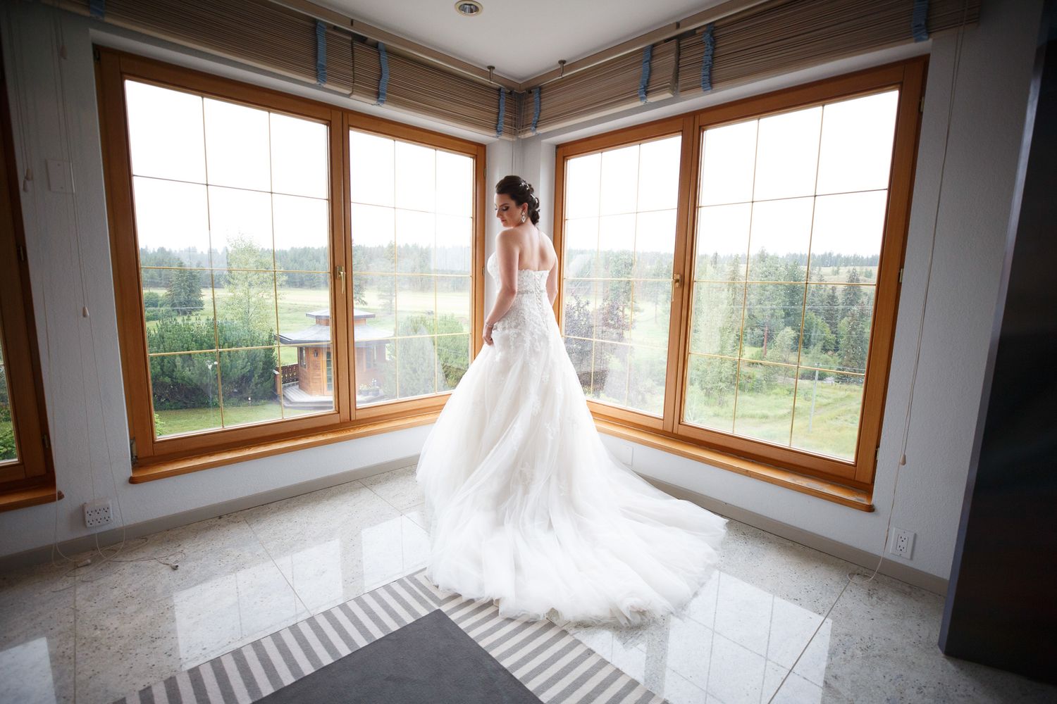 Bride in elegant white wedding gown with long train standing before large windows with scenic outdoor view at Cherry creek estate