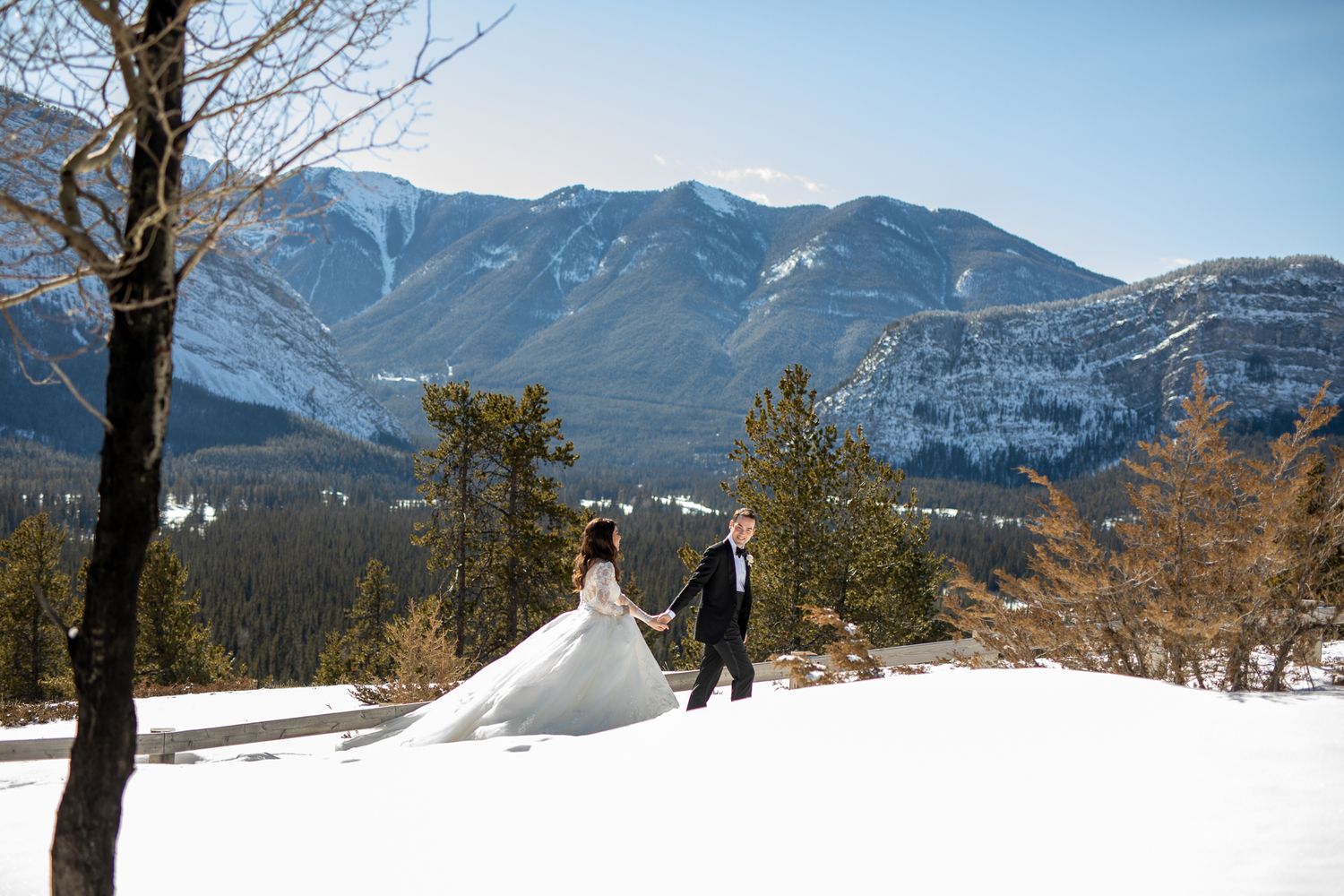 A couple in wedding attire walks through snowy terrain with majestic snow-capped mountains in the White Mountains backdrop in Banff, AB.