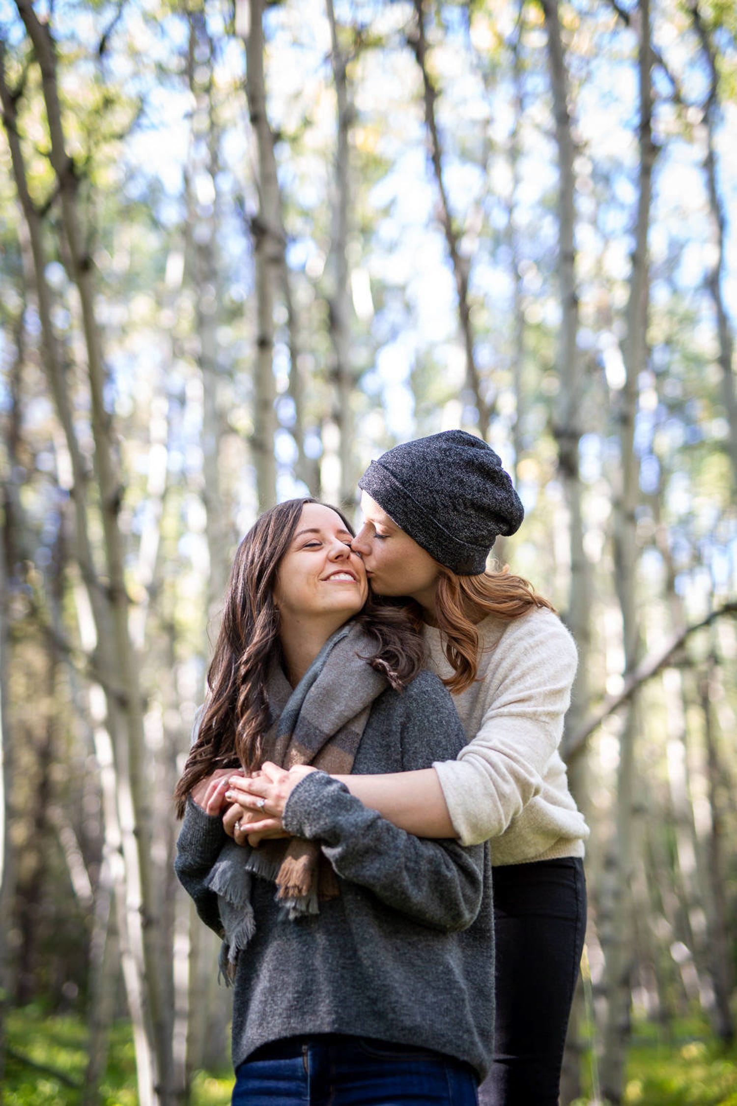 A LGBTQ couple embraces affectionately in a sunlit forest setting with tall trees in the background in Kananaskis Country in Alberta.