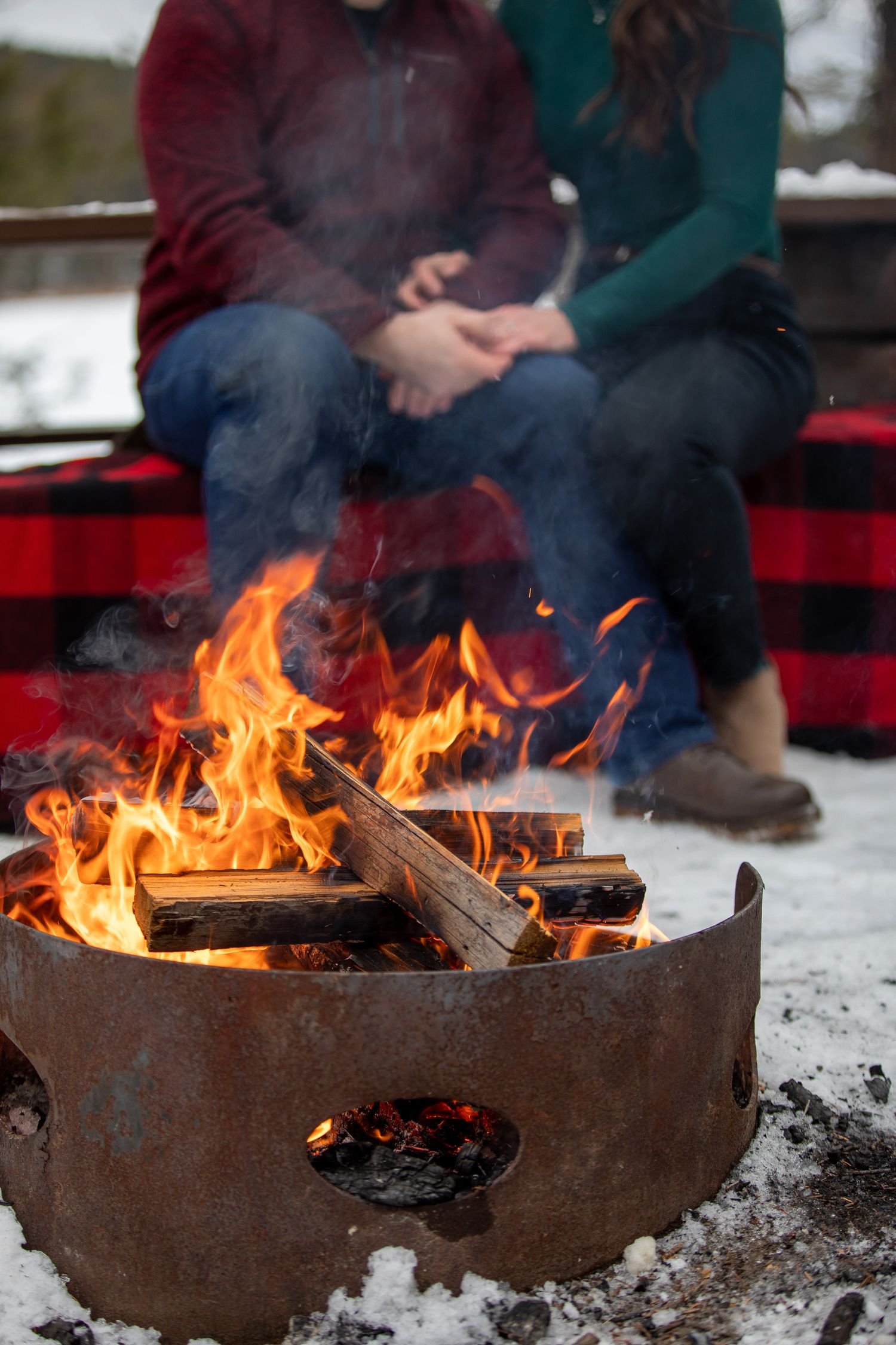 A rustic metal fire pit with roaring flames burns brightly in the snow during a winter gathering outdoors at Grey Wolf Panorama, BC