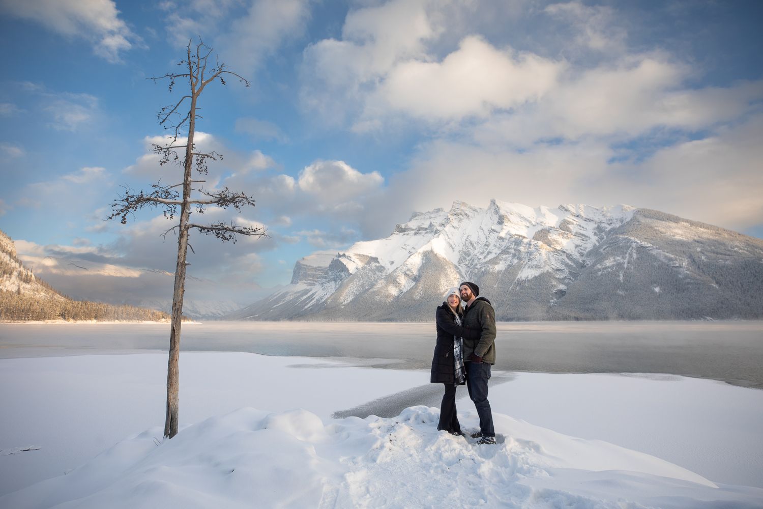A couple newly engaged standing on a rock in a wintery landscape of Minnewanka Lake in Banff, AB.